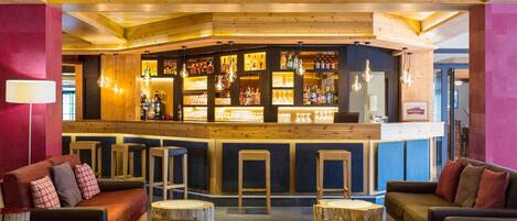 A bar area with wooden bar stools, a counter with bottles, and seating with round tables.