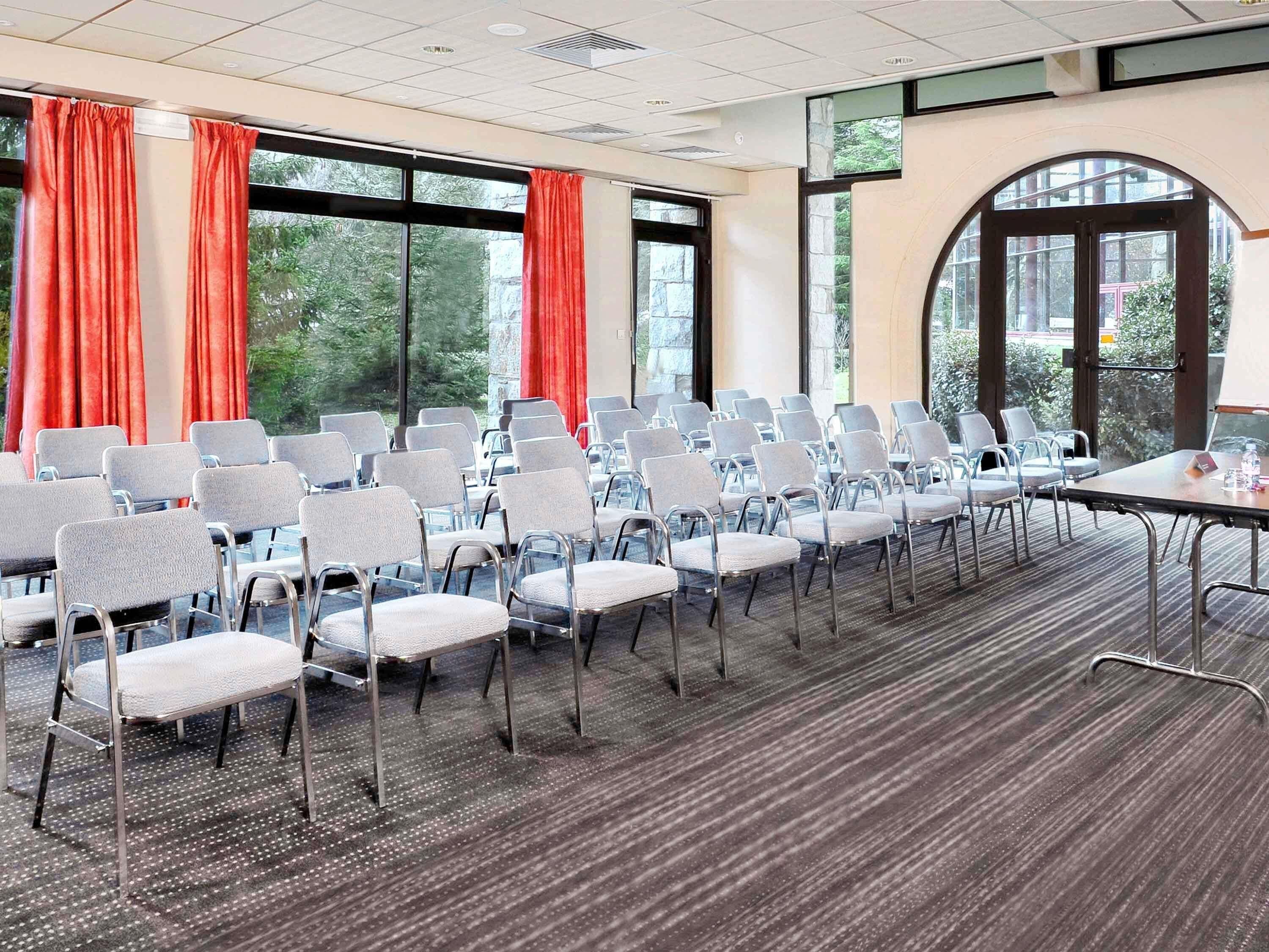 Un salón de conferencias con filas de sillas, una mesa y ventanas amplias con cortinas.