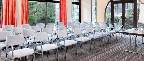 Un salón de conferencias con filas de sillas, una mesa y ventanas amplias con cortinas.