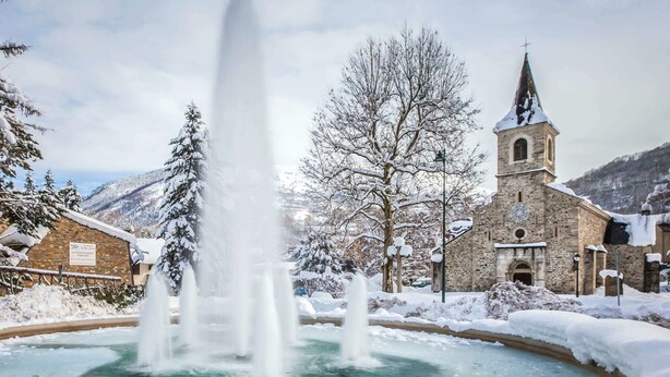 Un edificio de piedra histórico con una torre de iglesia, una fuente en primer plano y árboles y terreno cubiertos de nieve.