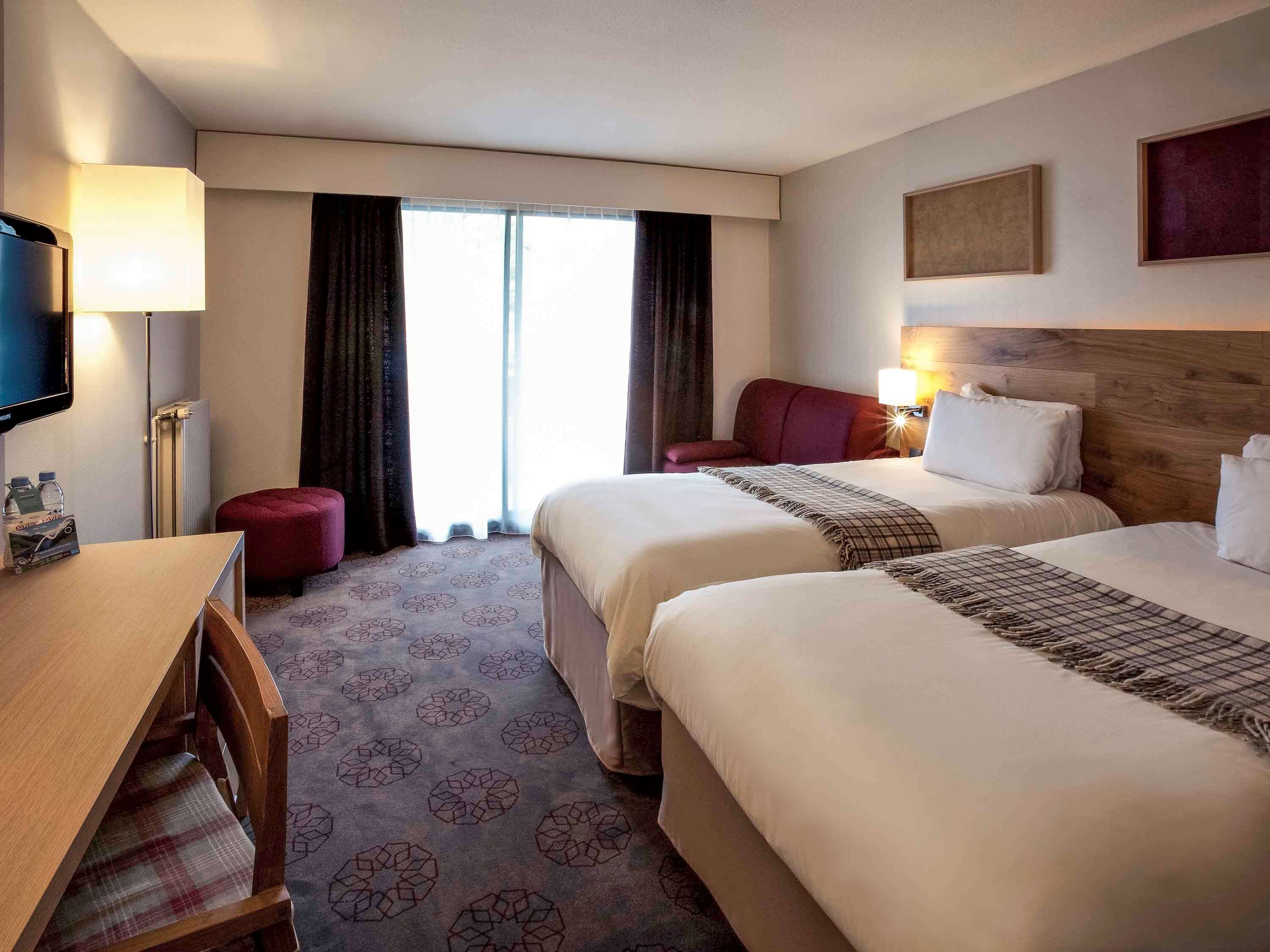 A hotel room with two beds, a desk, a TV, and a window with curtains.
