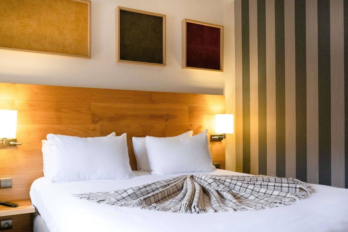Habitación de hotel con cabecera de madera, dos lámparas de noche, cama con ropa de cama blanca y una manta de cuadros.