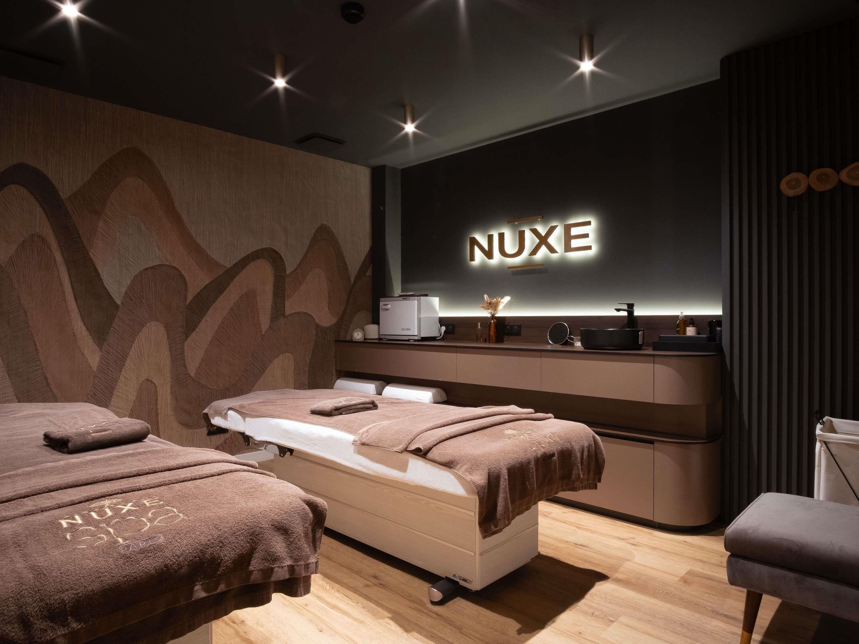 A modern spa room with a massage table, two towels, a reception desk, and a wall-mounted sign reading 'NUXE'.