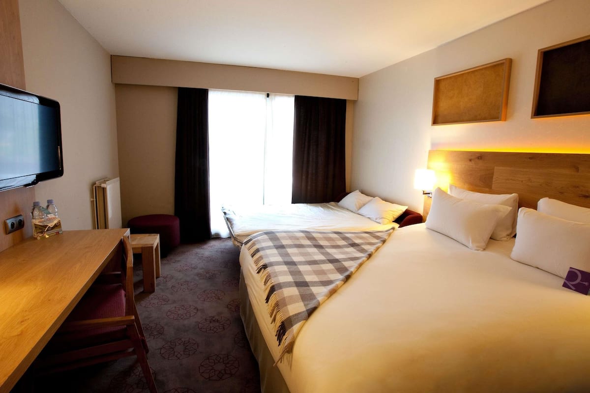 Una habitación de hotel con cama, escritorio, televisor, ventana con cortinas y lámpara.