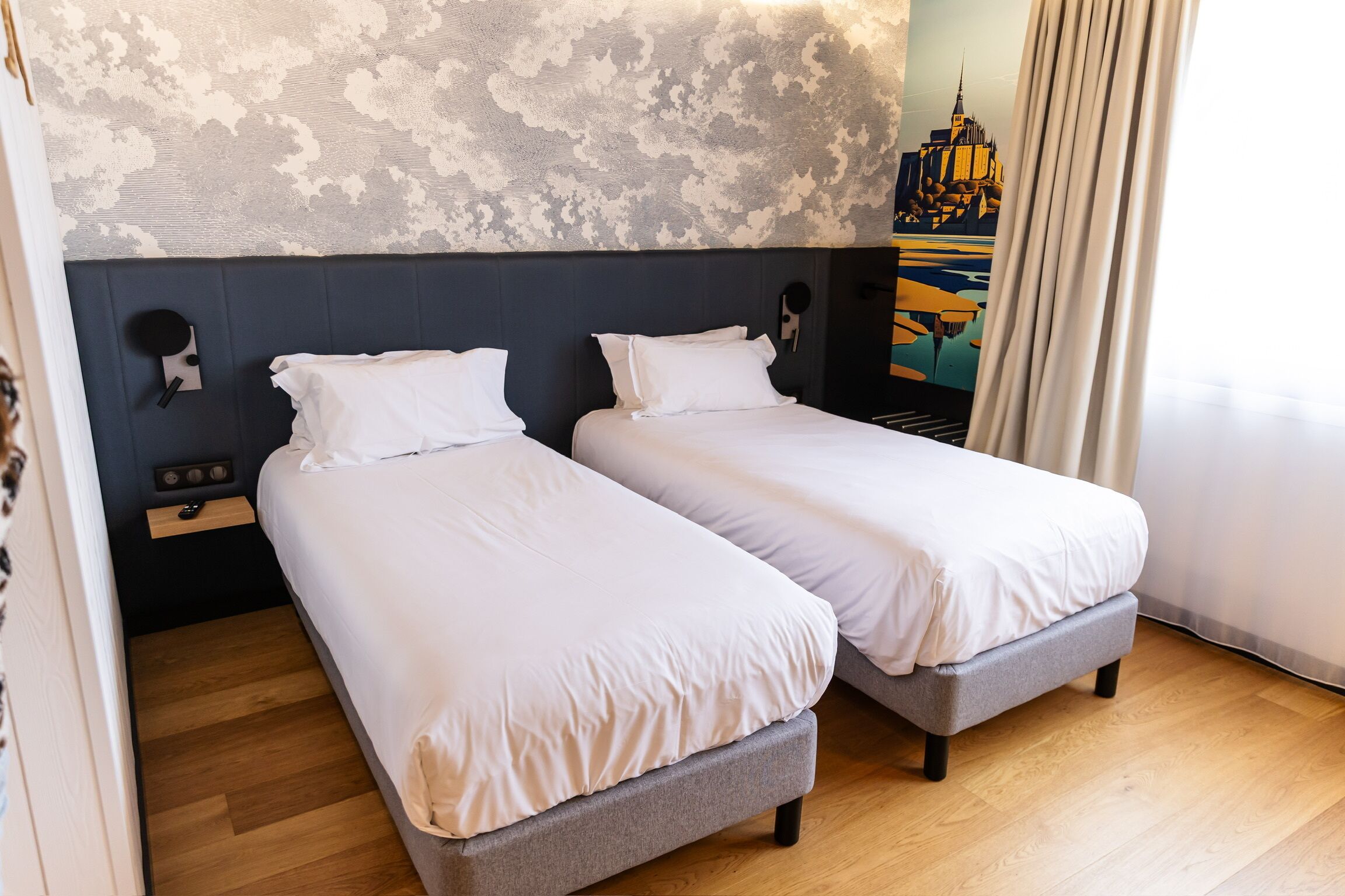 Ein Hotelzimmer mit zwei Einzelbetten, Holzboden, einem gemusterten Wandbild und einem Stadtbildgemälde.