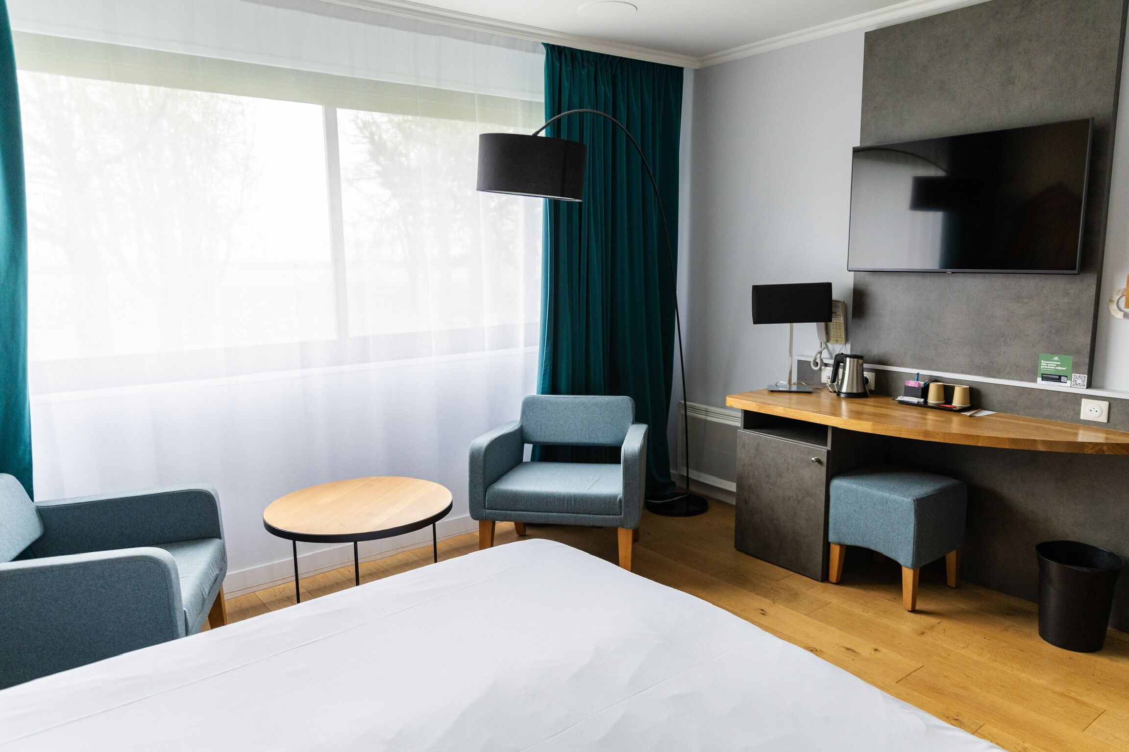 A hotel room with a bed, a desk, a chair, a small table, and a trash bin.