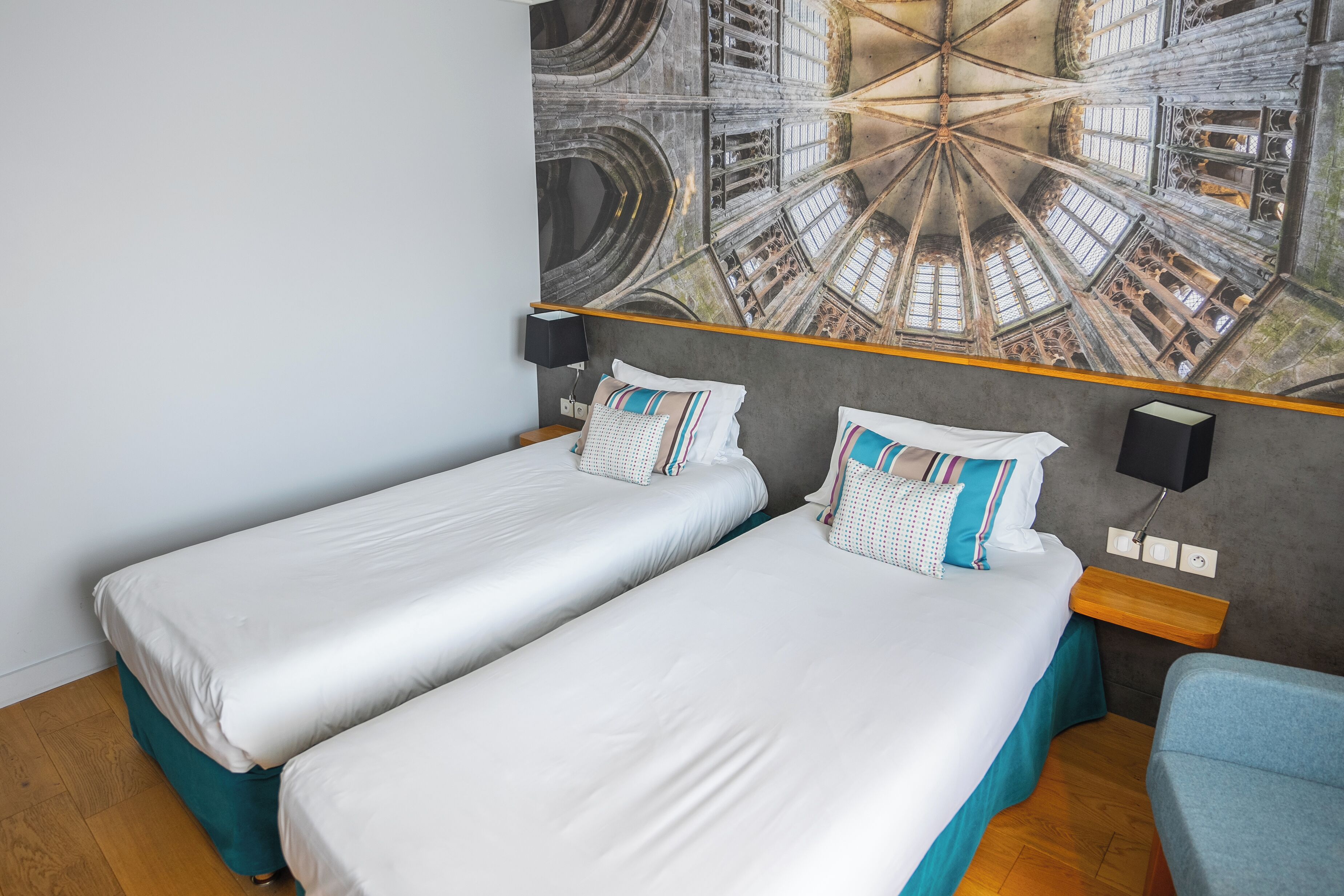 A hotel room with two single beds, a blue chair, a wall tapestry, and a wooden floor.