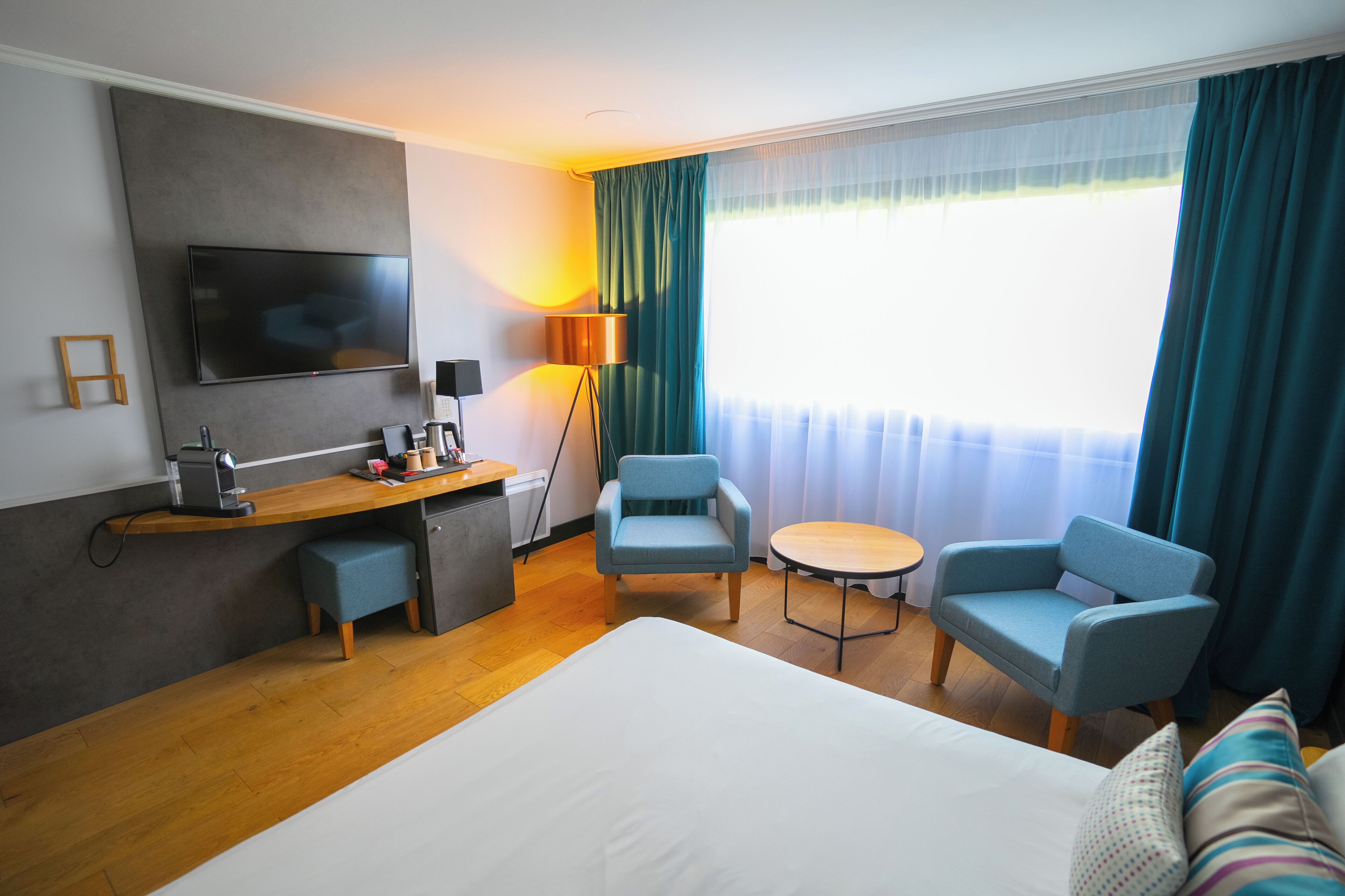 A hotel room with a bed, a desk, a chair, a small table, a TV, and a lamp.