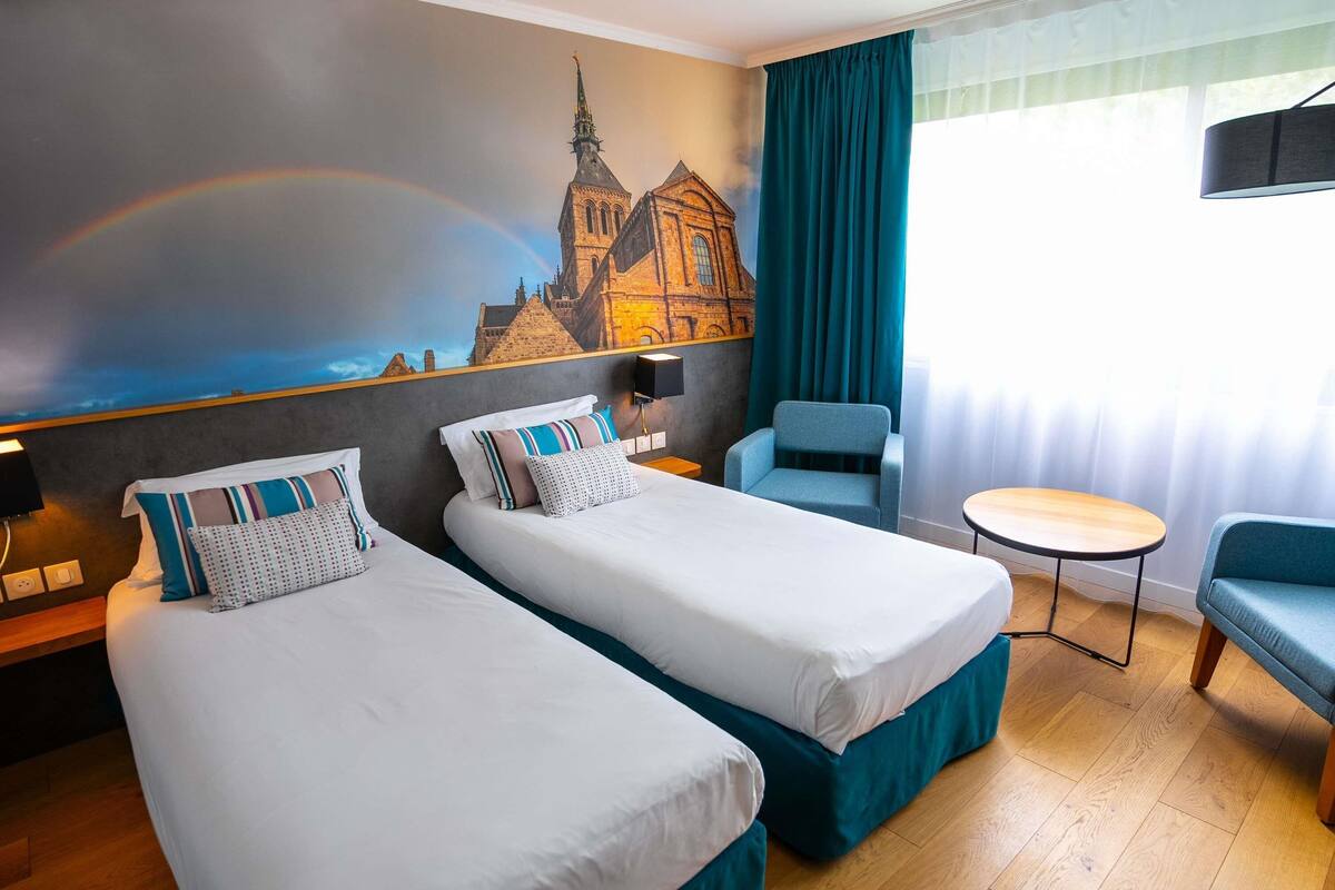 Une chambre d’hôtel avec deux lits, une chaise, une petite table et une fresque représentant une église.