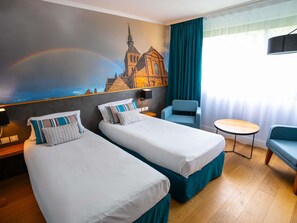 Ein Hotelzimmer mit zwei Betten, einem Sessel, einem kleinen Tisch und einem Wandgemälde einer Kirche.
