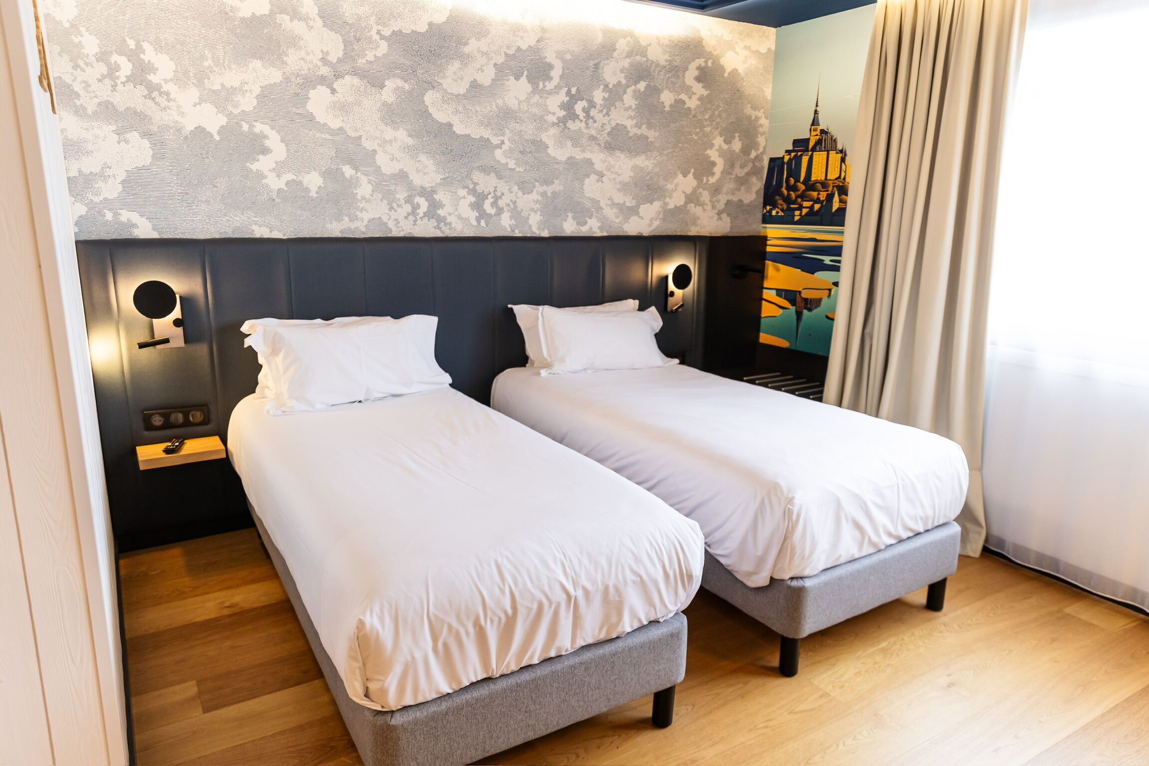 Una habitación de hotel con dos camas individuales, suelo de madera, pared con diseño y un cuadro de un paisaje urbano.