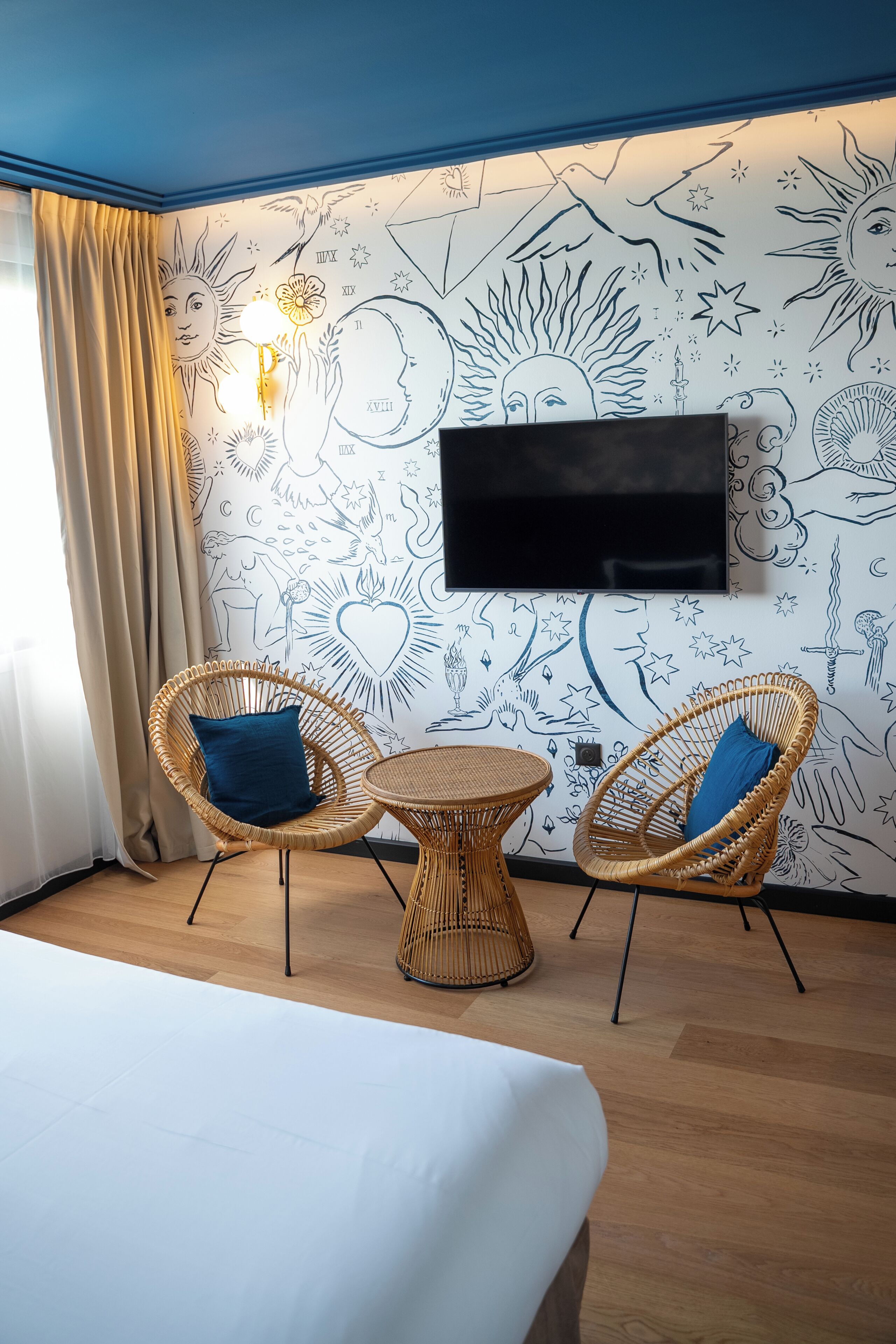 A hotel room with a bed, two wicker chairs, a small round table, and a wall-mounted TV.