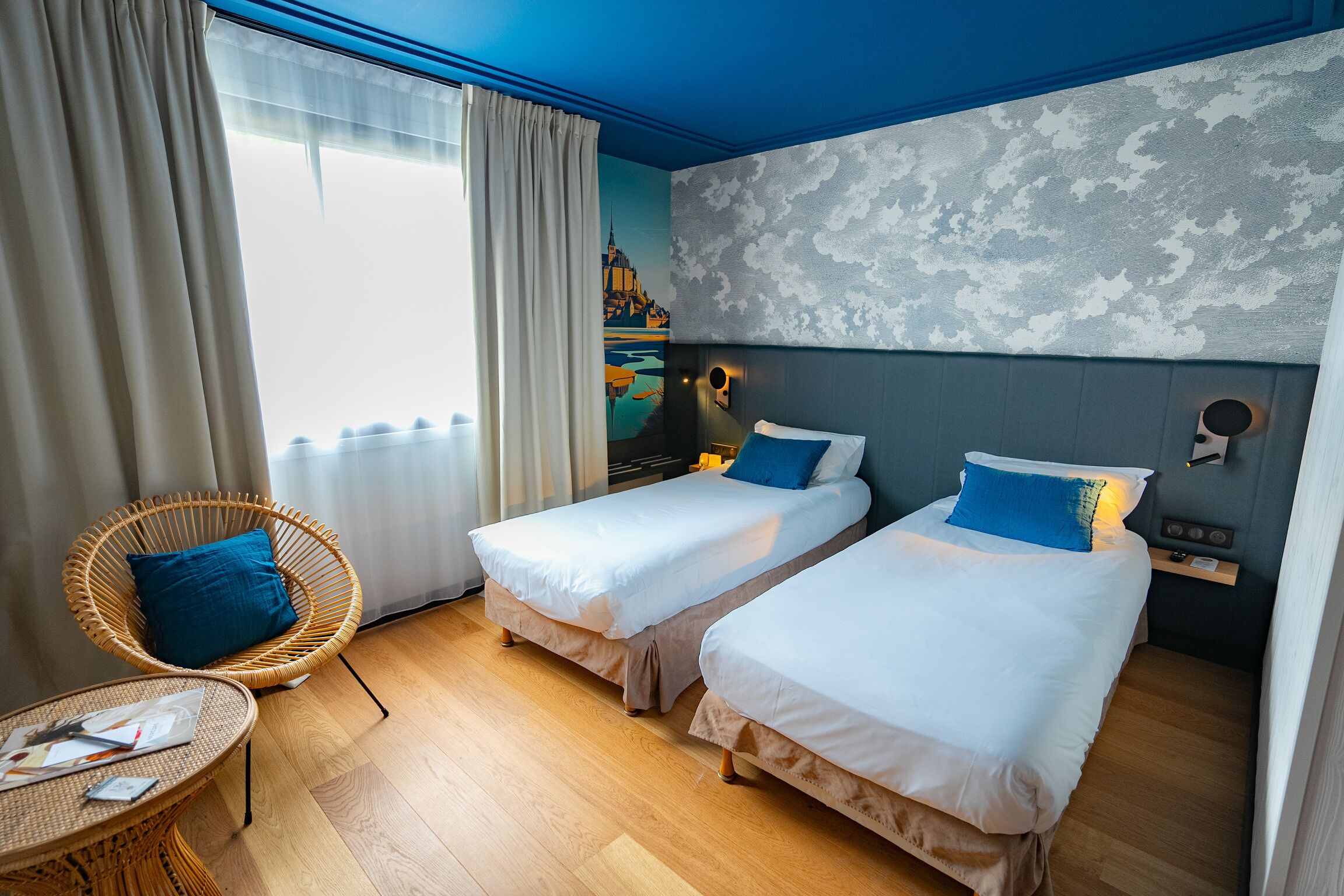 Quarto de hotel com duas camas individuais, uma cadeira de vime, uma mesa pequena e vista para a paisagem urbana pela janela.