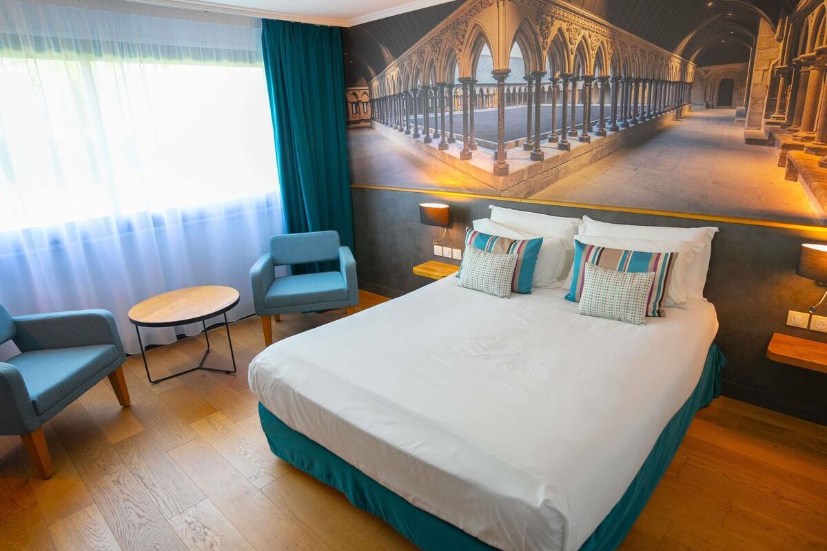 Une chambre d’hôtel avec un grand lit, une petite table ronde, un fauteuil bleu et une fresque représentant l’intérieur d’une cathédrale.