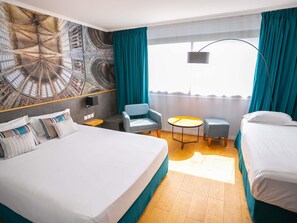 Ein Hotelzimmer mit einem großen Bett, zwei Einzelbetten, einem Schreibtisch, einem Sessel, einem kleinen Tisch und einem Wandgemälde.