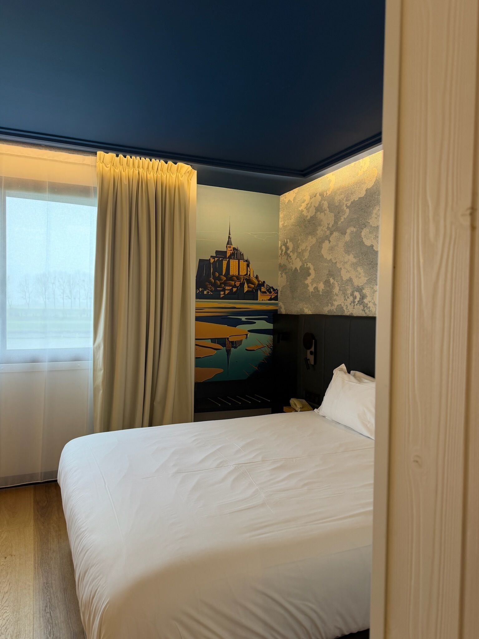 Una habitación de hotel con una cama, un ventanal con cortinas, un cuadro en la pared y un papel tapiz estampado.