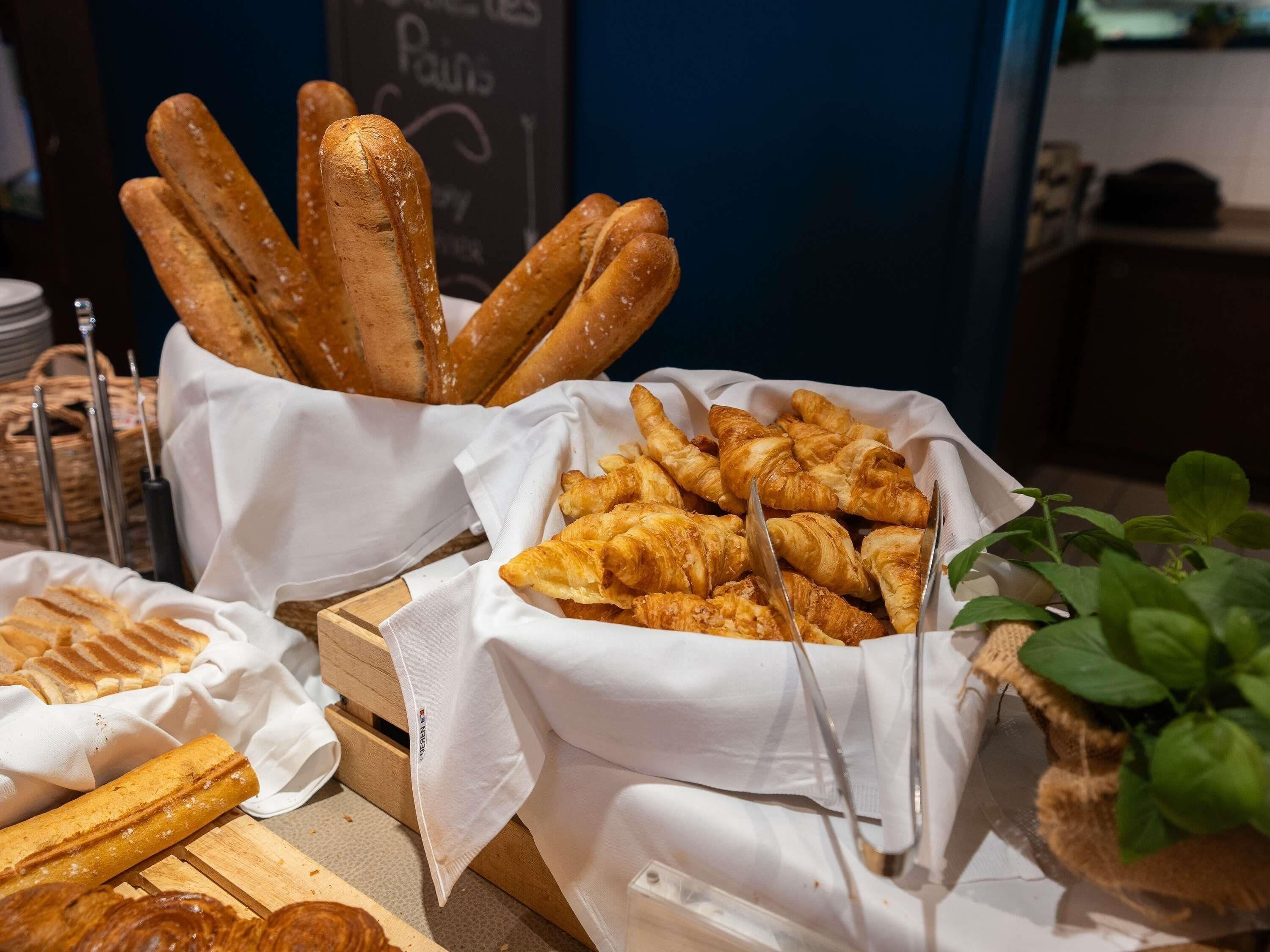 A display of bread and pastries, including baguettes and croissants, arranged on a wooden table with white cloths.