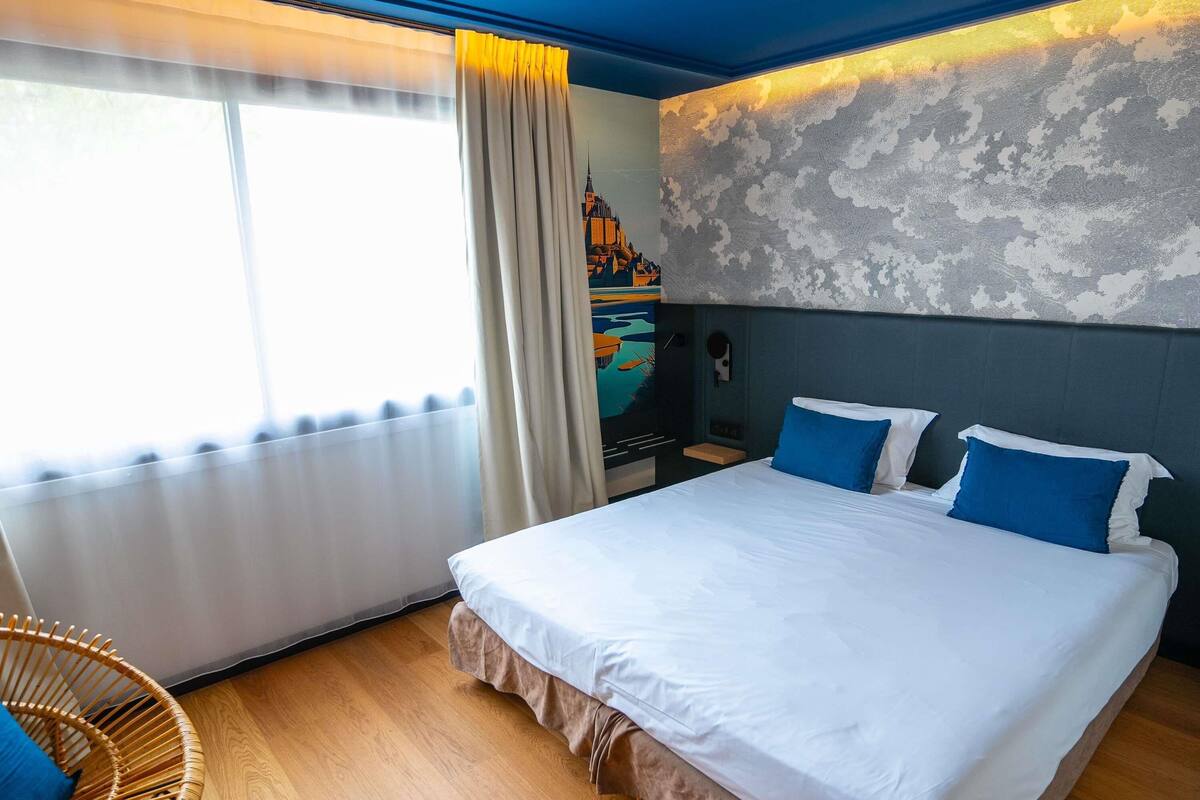 Une chambre d’hôtel avec un grand lit, une chaise, un tableau au mur et une fenêtre avec des rideaux.
