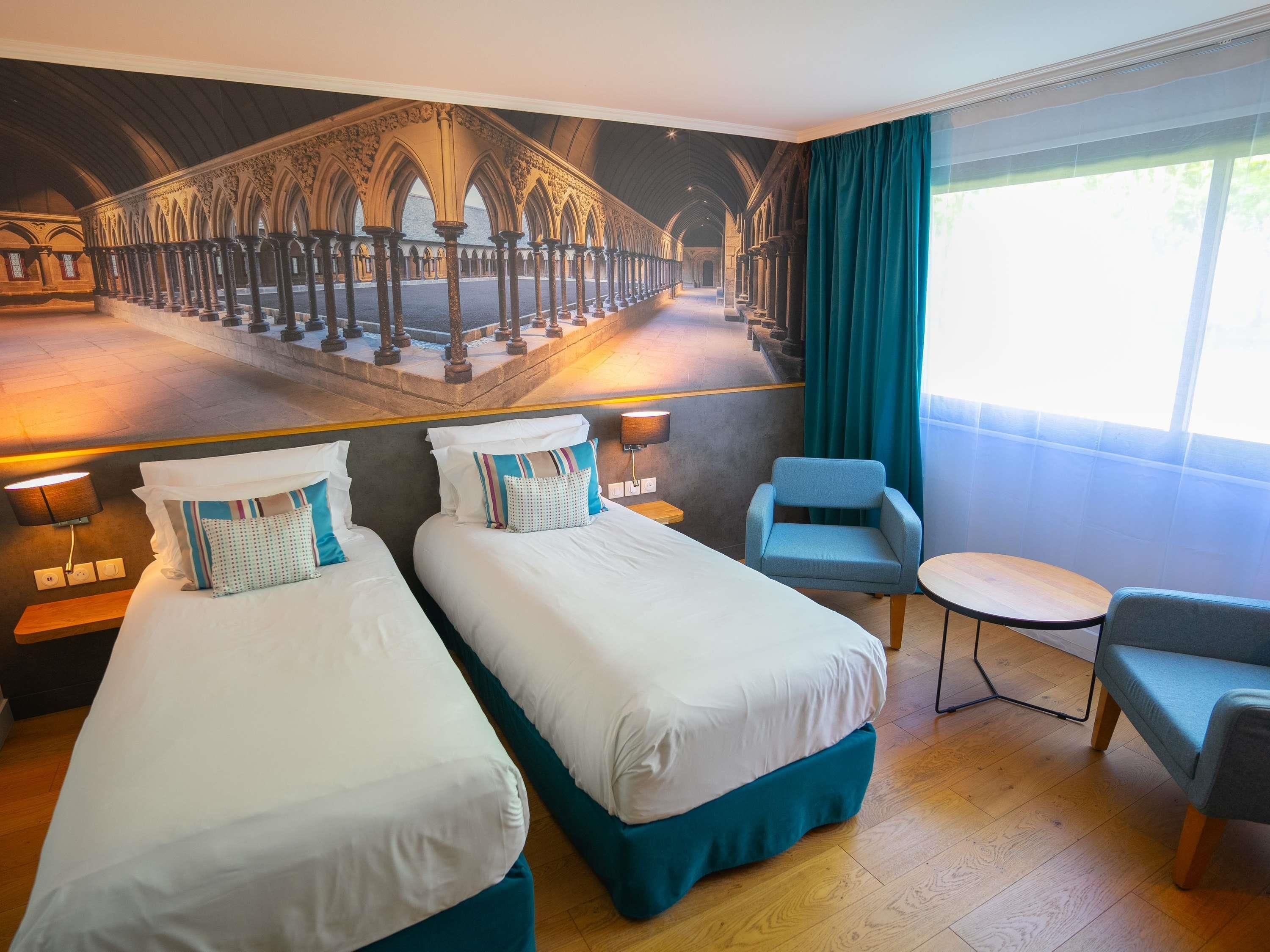 A hotel room with two beds, a small table, a chair, and a large window with curtains.