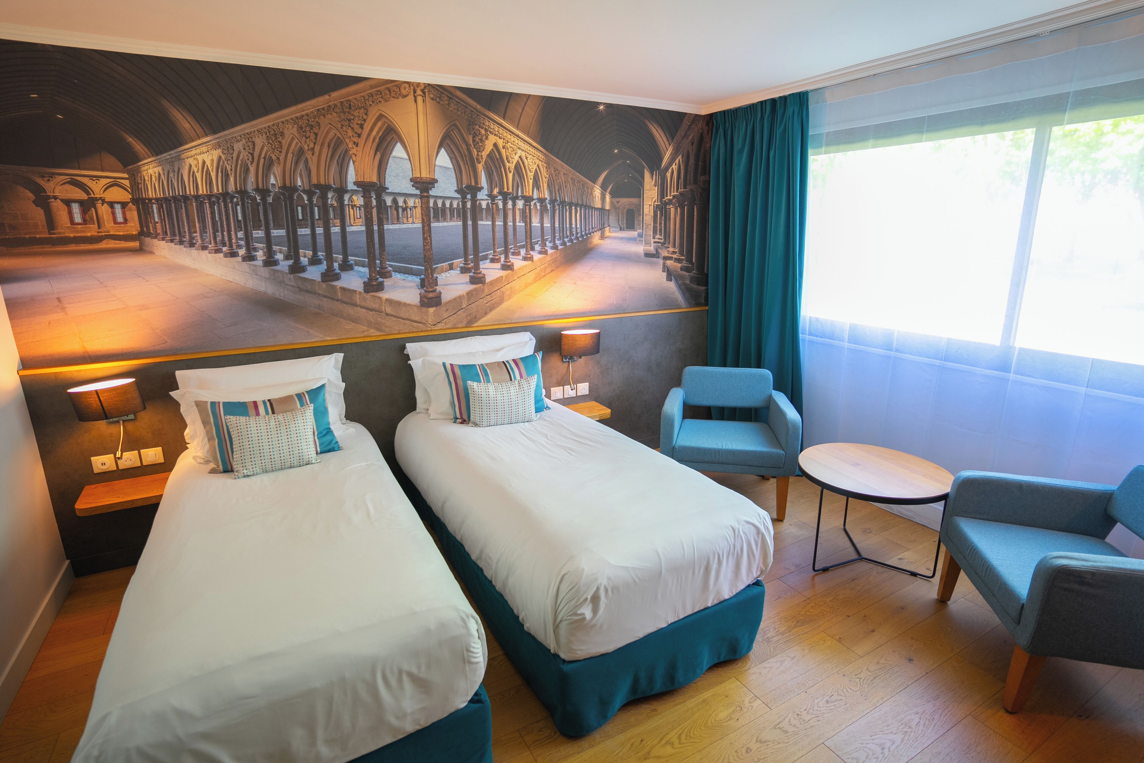 A hotel room with two beds, a small table, a chair, and a view of a cathedral.