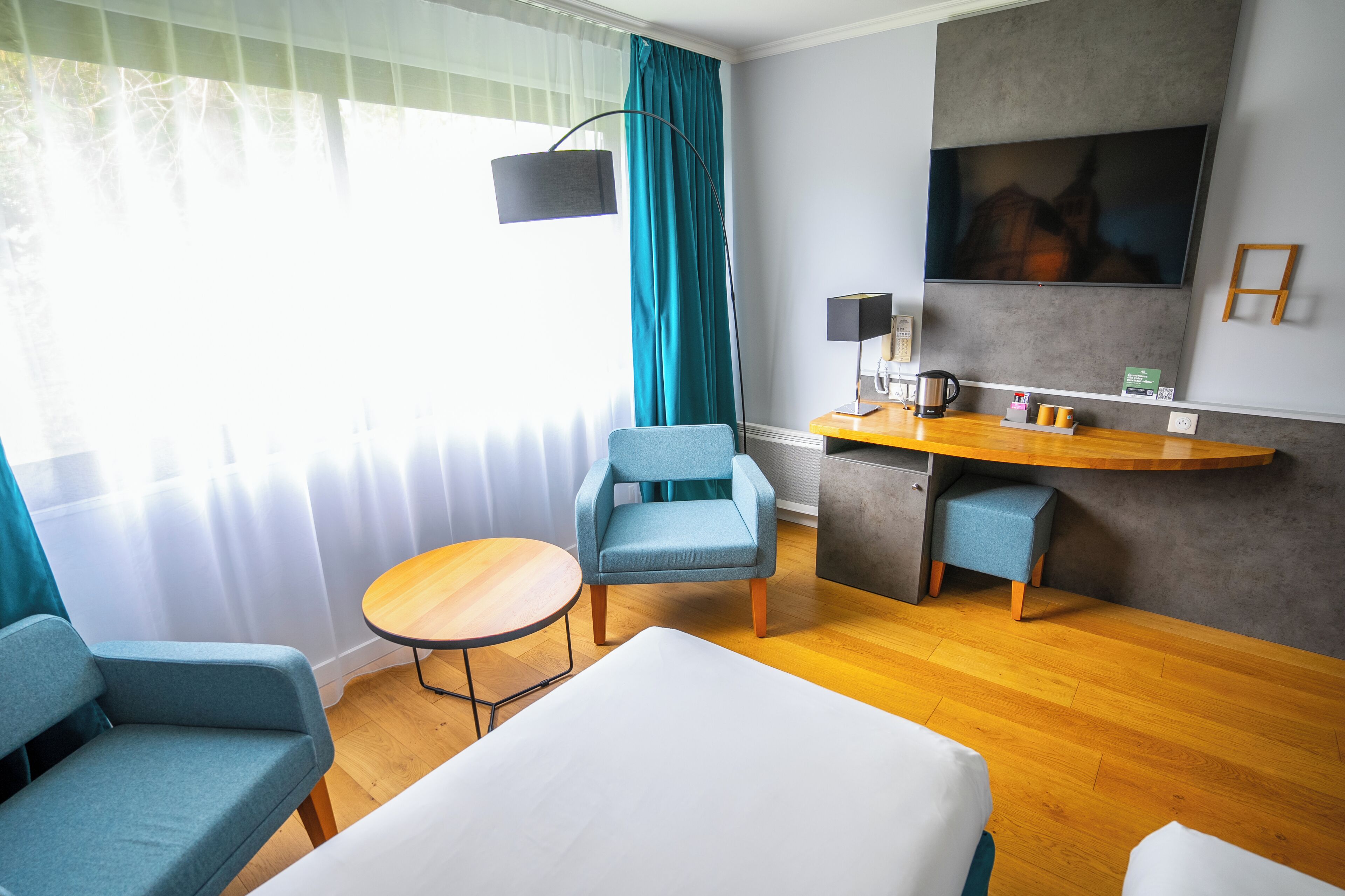 Ett hotellrum med en säng, två fåtöljer, ett litet bord, ett skrivbord med en lampa, en kaffemaskin och en tv på väggen.