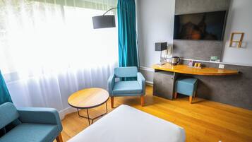 Ett hotellrum med en säng, två fåtöljer, ett litet bord, ett skrivbord med en lampa, en kaffemaskin och en tv på väggen.
