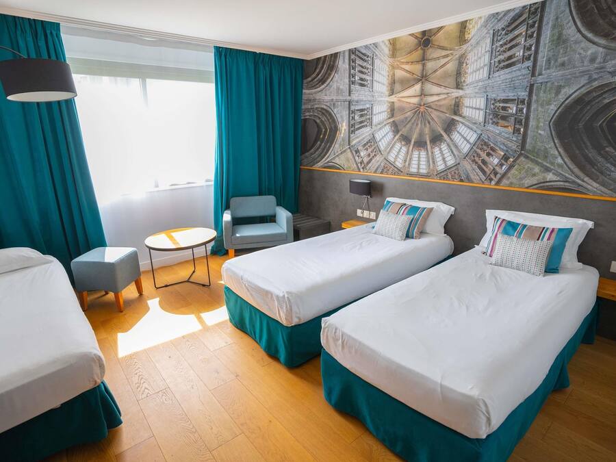 A hotel room with two single beds, a small table, a chair, and a large window with curtains.