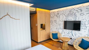 Une chambre d’hôtel moderne comprenant un lit, deux fauteuils en osier, une petite table, un téléviseur fixé au mur et un mur décoratif orné de motifs abstraits.