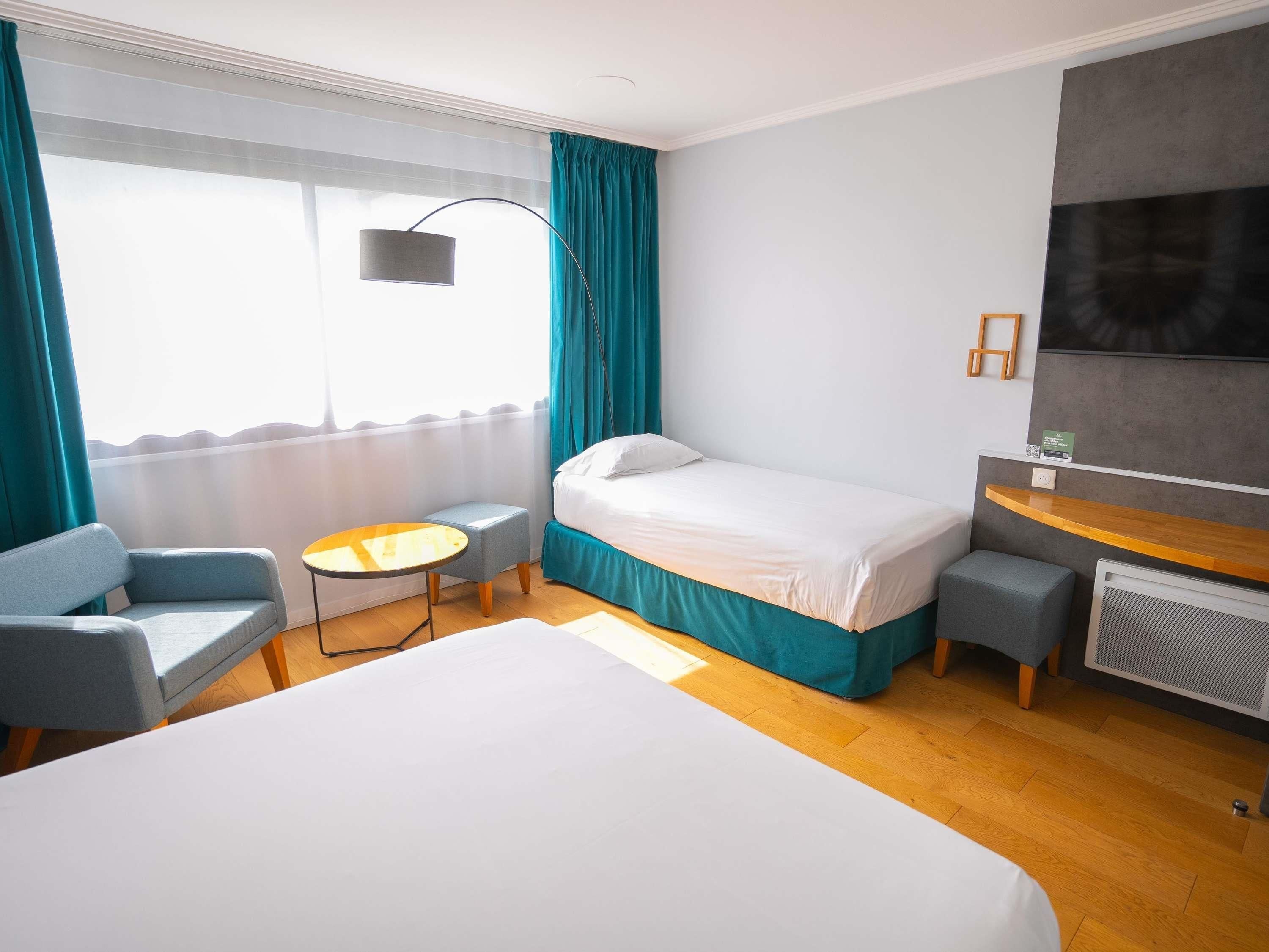 A hotel room with a bed, a desk, a chair, a small table, and a large window with curtains.