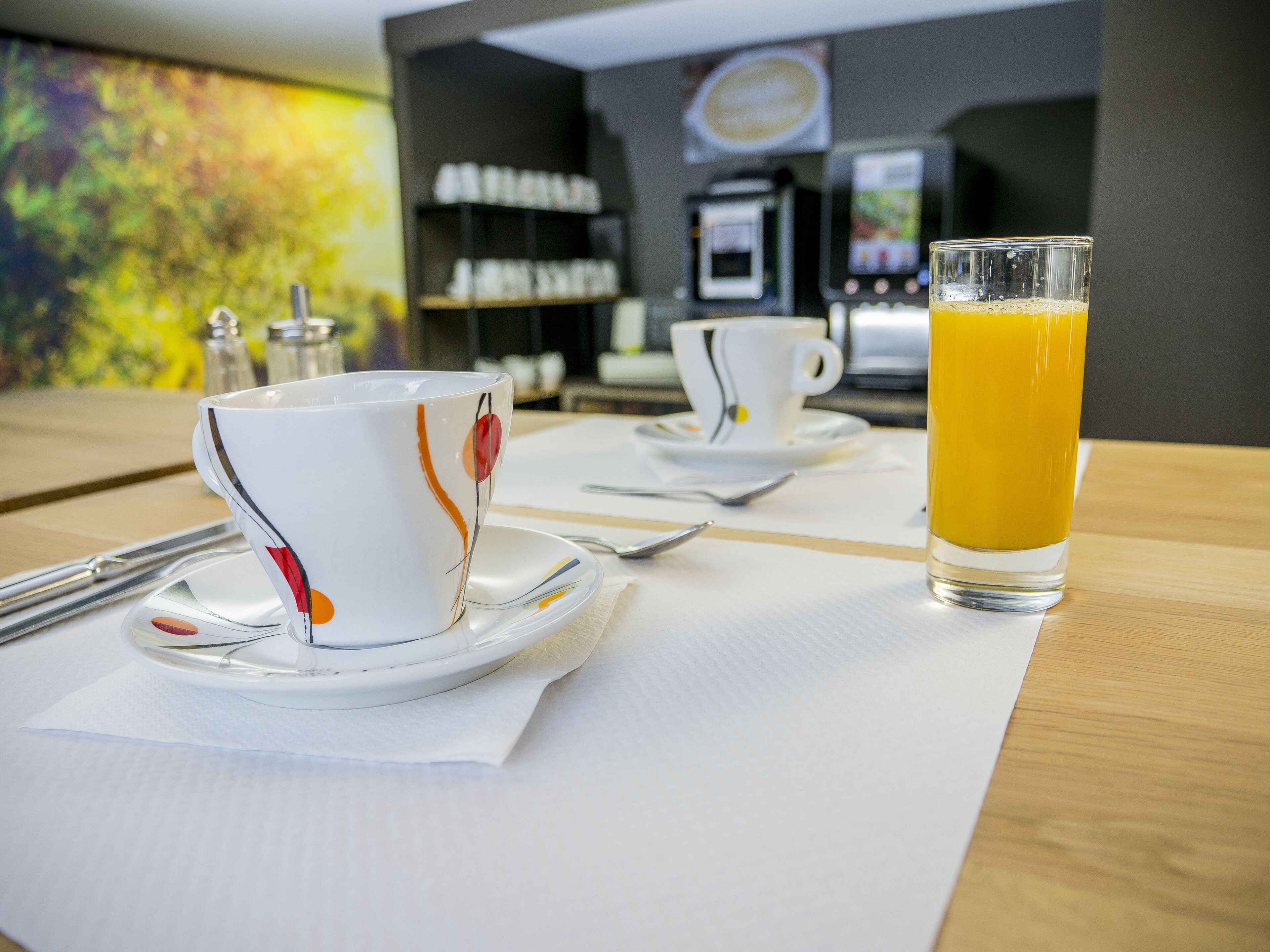 A breakfast setting with a table set for two, including a glass of orange juice and two cups with saucers.