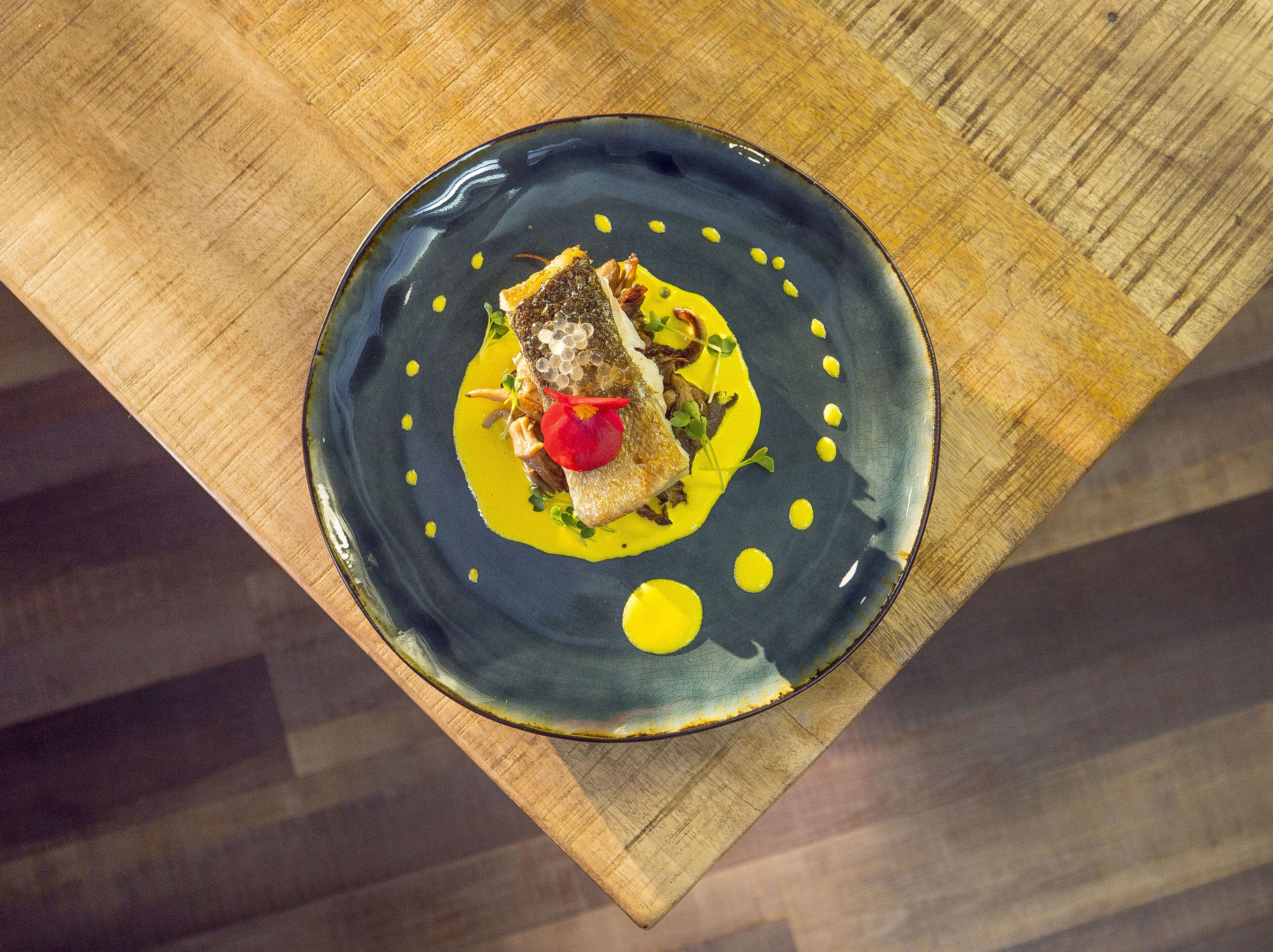Une assiette en céramique sombre sur laquelle est présenté un plat de poisson, le tout posé sur une table en bois.