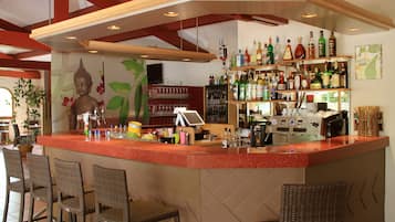 Zona de bar con una estatua de Buda, taburetes, una barra con una cafetera y estantes con diversas botellas.