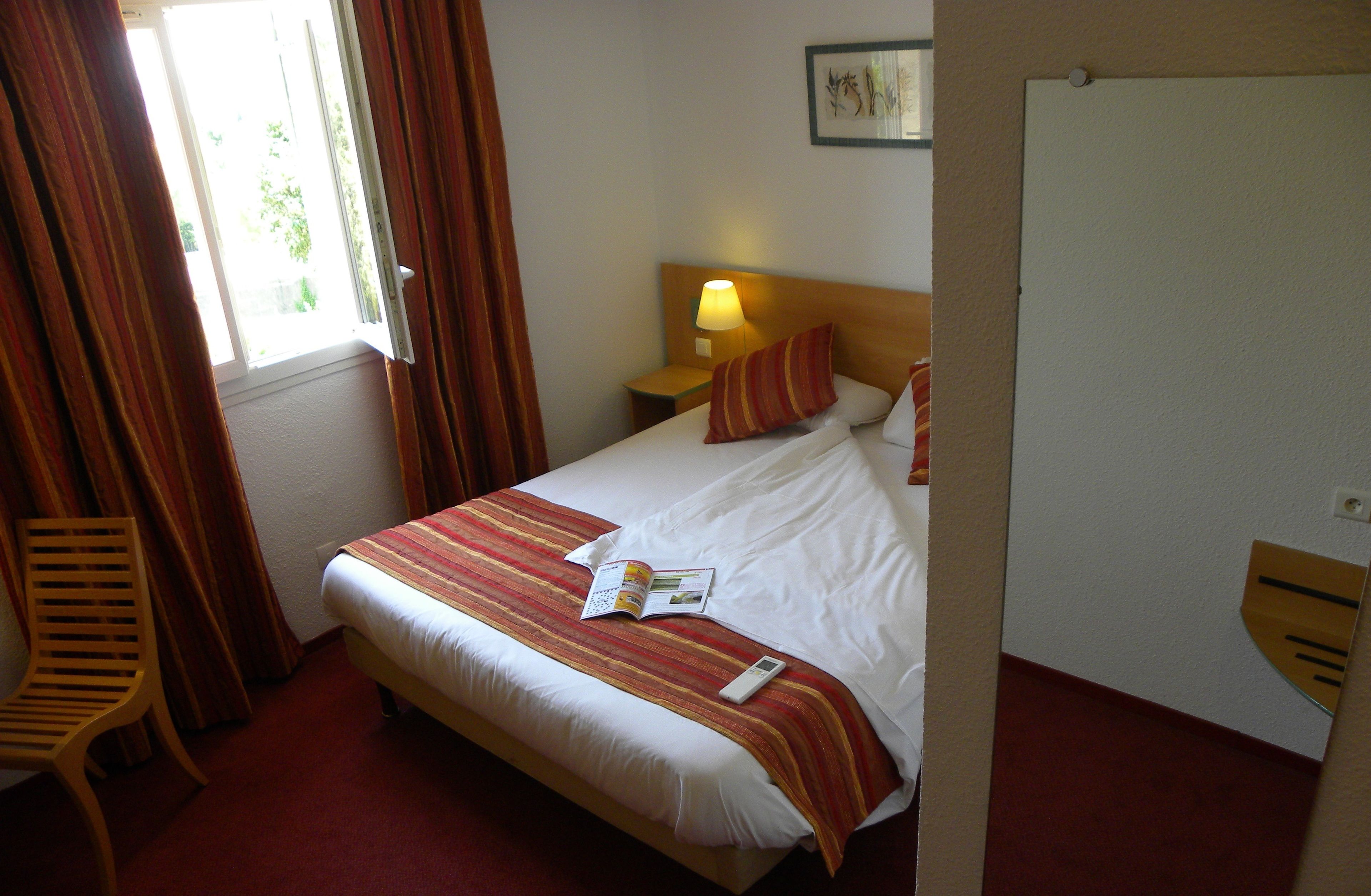 Une chambre d’hôtel avec un lit, une table de chevet avec une lampe, une chaise et une fenêtre avec des rideaux.