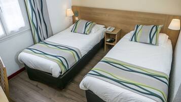 A hotel room with two single beds, each with striped bedding, a wooden headboard, bedside lamps, a small bedside table with a phone, and a window with curtains.