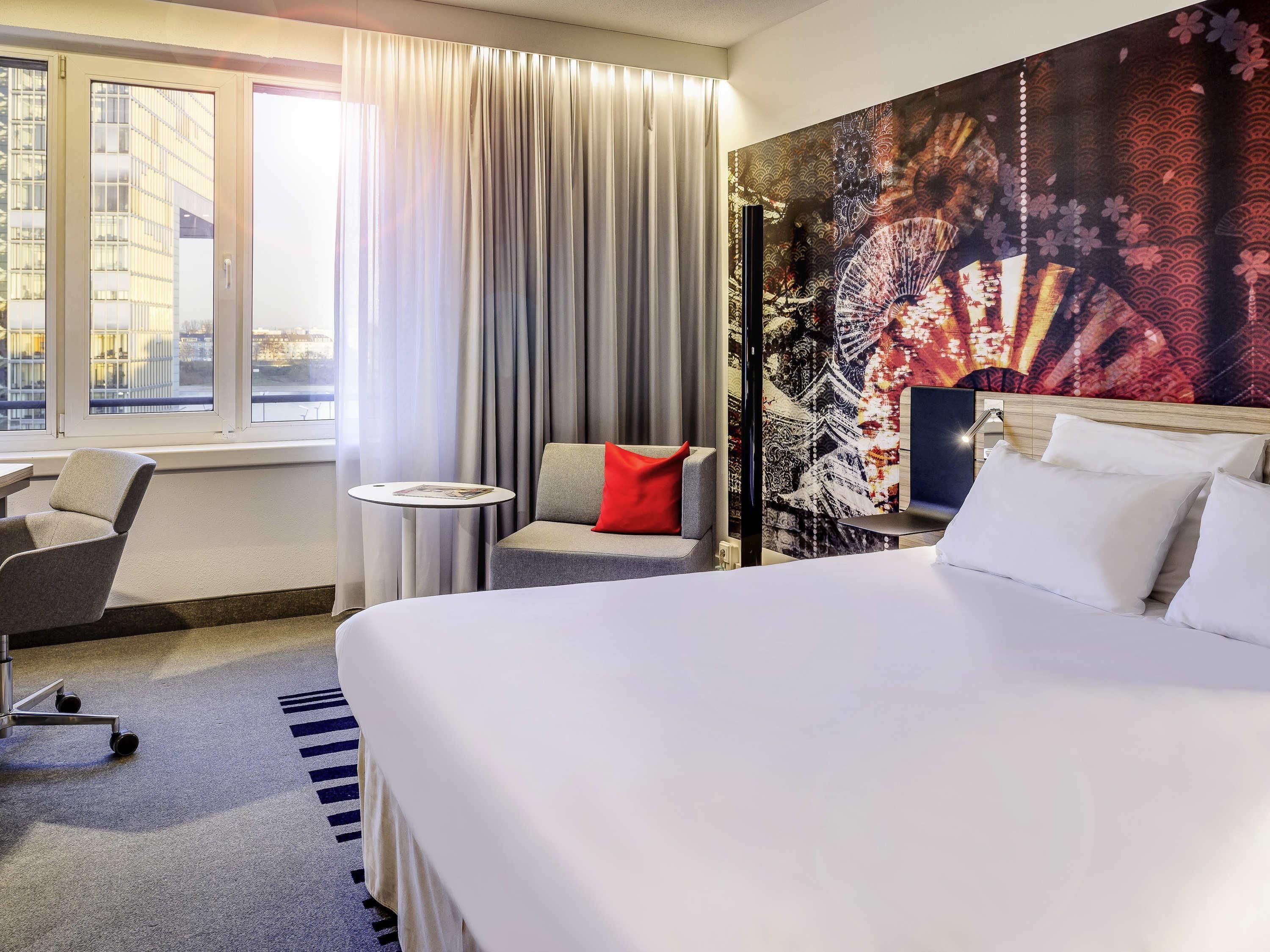 Quarto de hotel com cama, uma cadeira, uma mesinha e uma janela grande.