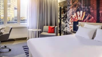 Quarto de hotel com cama, uma cadeira, uma mesinha e uma janela grande.