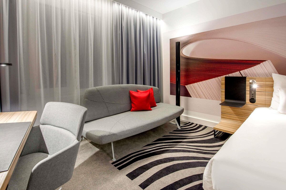 A modern hotel room with a grey sofa, a red pillow, a striped rug, and a bed with a wooden headboard.