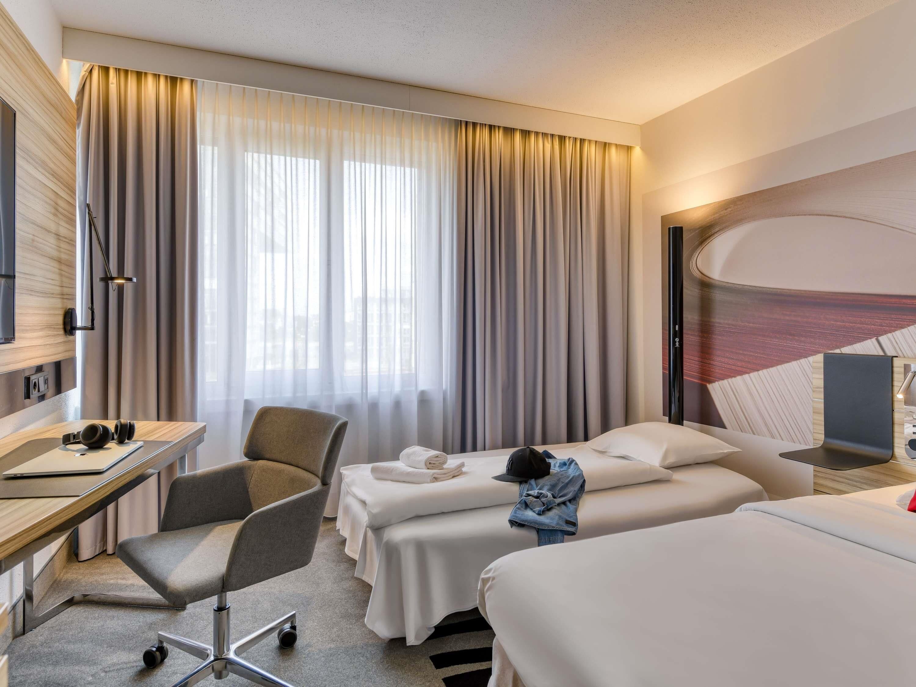 A hotel room with a desk, two beds, a chair, and a large window with curtains.