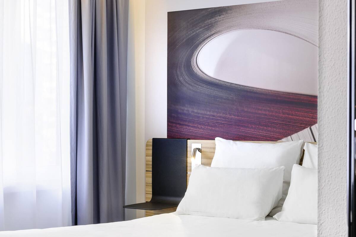 Hotellihuone, jossa on sänky, yöpöytä, lamppu ja suuri, abstrakti taulu seinällä.