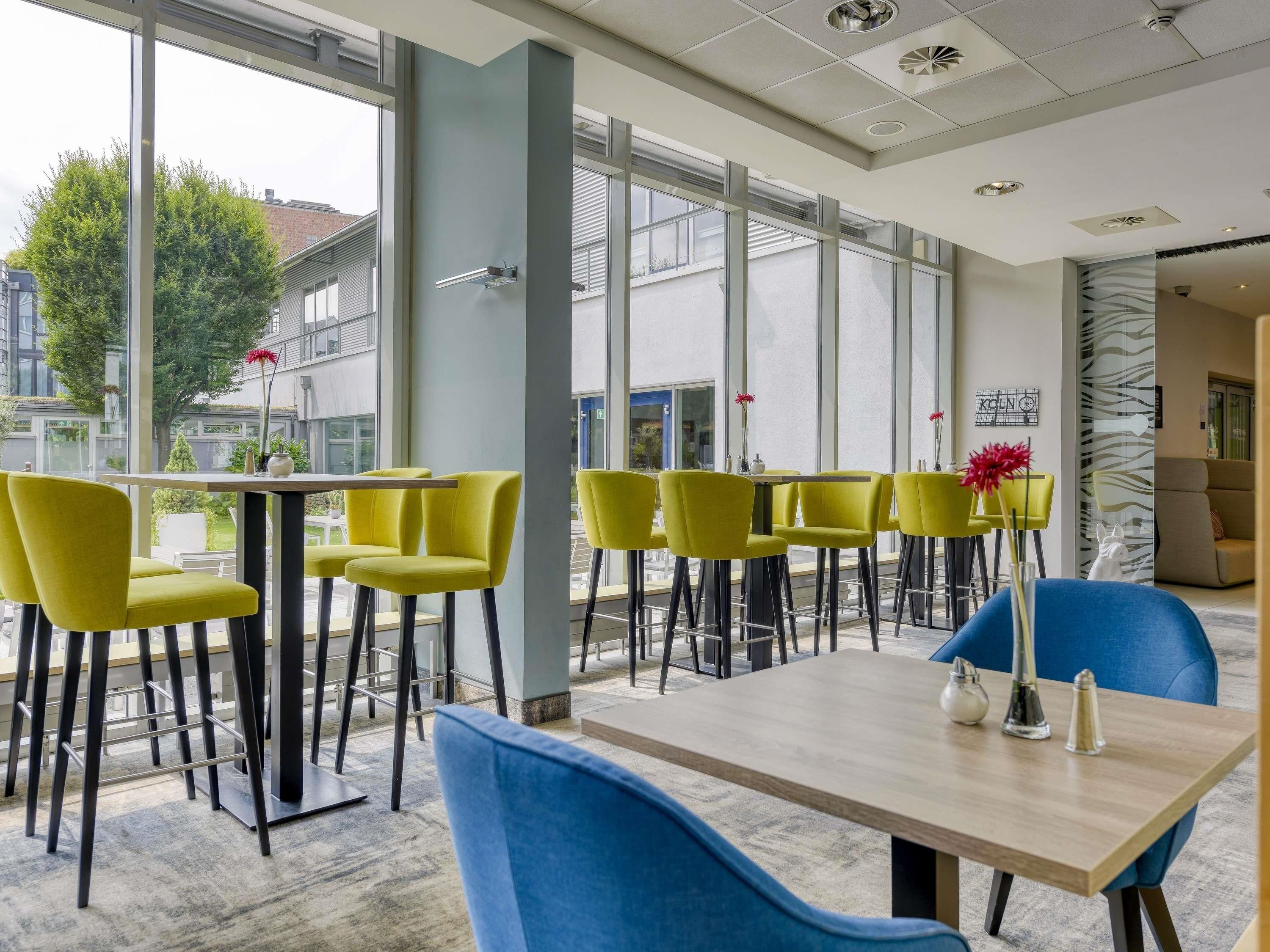 A modern interior with yellow chairs, a wooden table, and large windows offering views of buildings and trees.