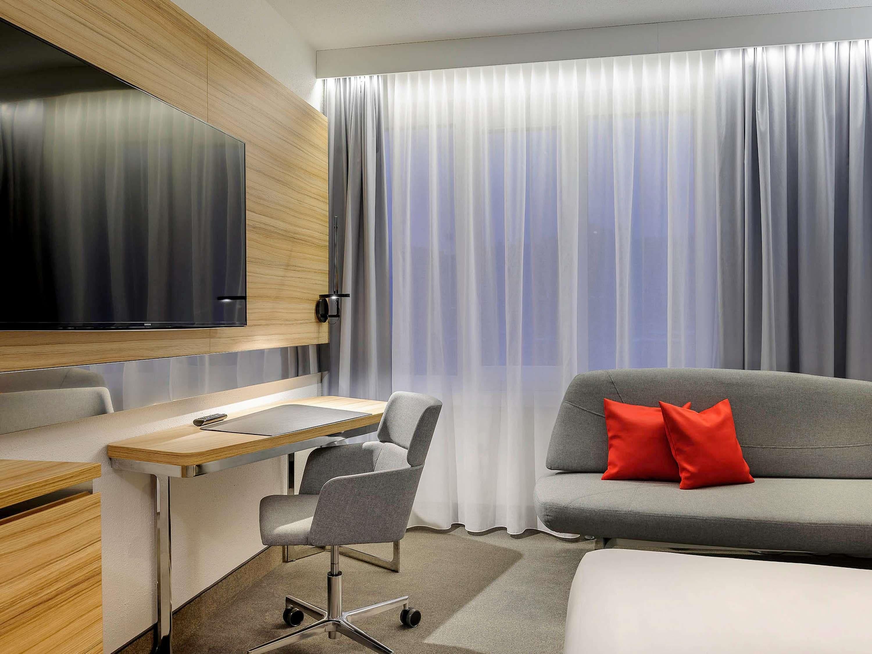 Et moderne hotelværelse med skrivebord, stol, sofa og et stort vindue med gardiner.