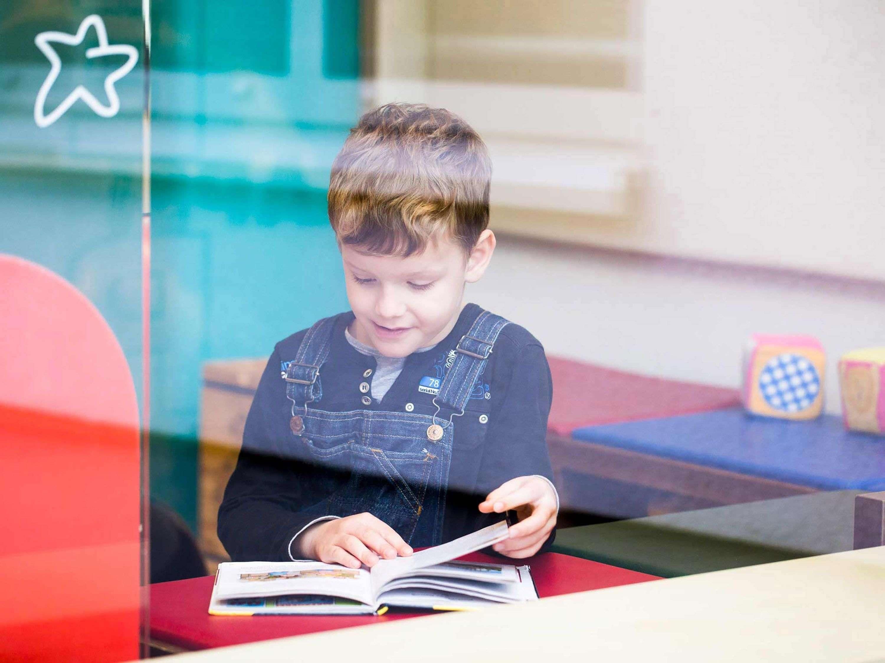 Un niño leyendo un libro en una mesa con mantel rojo, una decoración de estrella en la ventana y bloques de colores en el suelo.