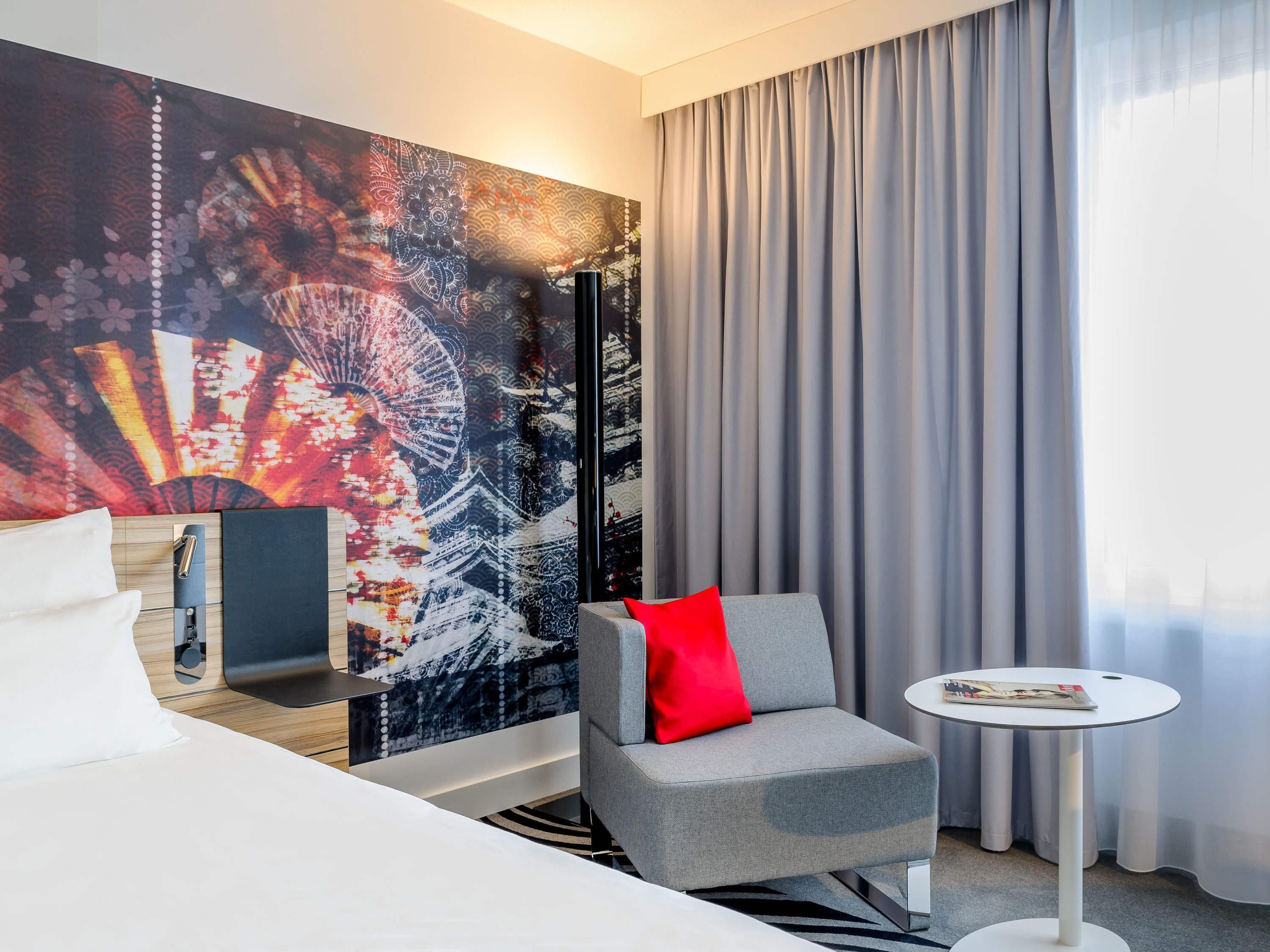 Una habitación de hotel moderna con un cuadro abstracto grande en la pared, un sillón gris con un cojín rojo, una mesita redonda pequeña y una cama con ropa de cama blanca.