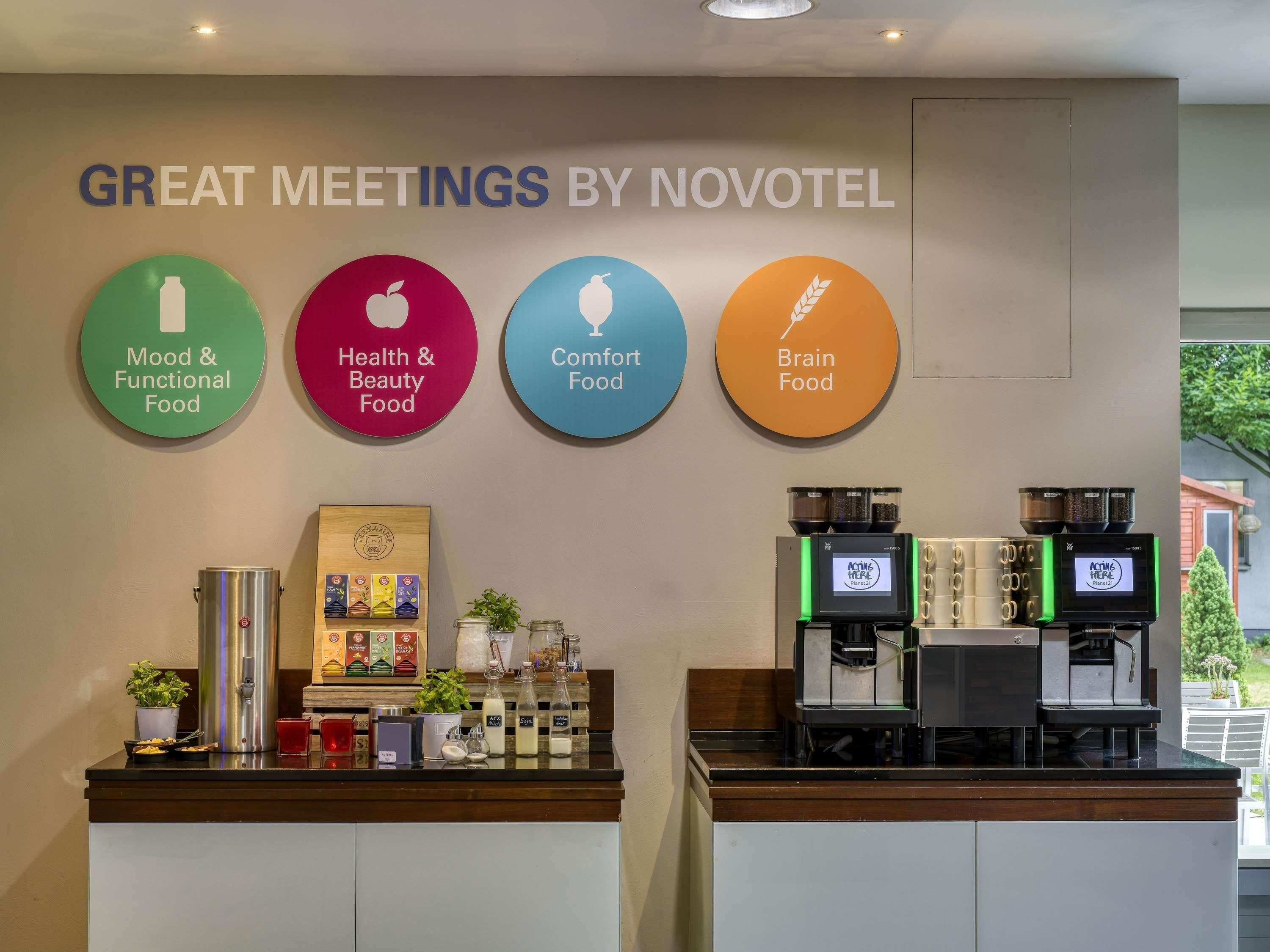 Ein Hotel-Empfangsbereich mit einem Schild, das 'GREAT MEETINGS BY NOVOTEL' anzeigt, und vier runde Schilder mit den Aufschriften 'Mood & Functional Food', 'Health & Beauty Food', 'Comfort Food' und 'Brain Food'. Zwei Kaffeemaschinen und eine Produktpräsentation von Lebensmitteln sind ebenfalls vorhanden.