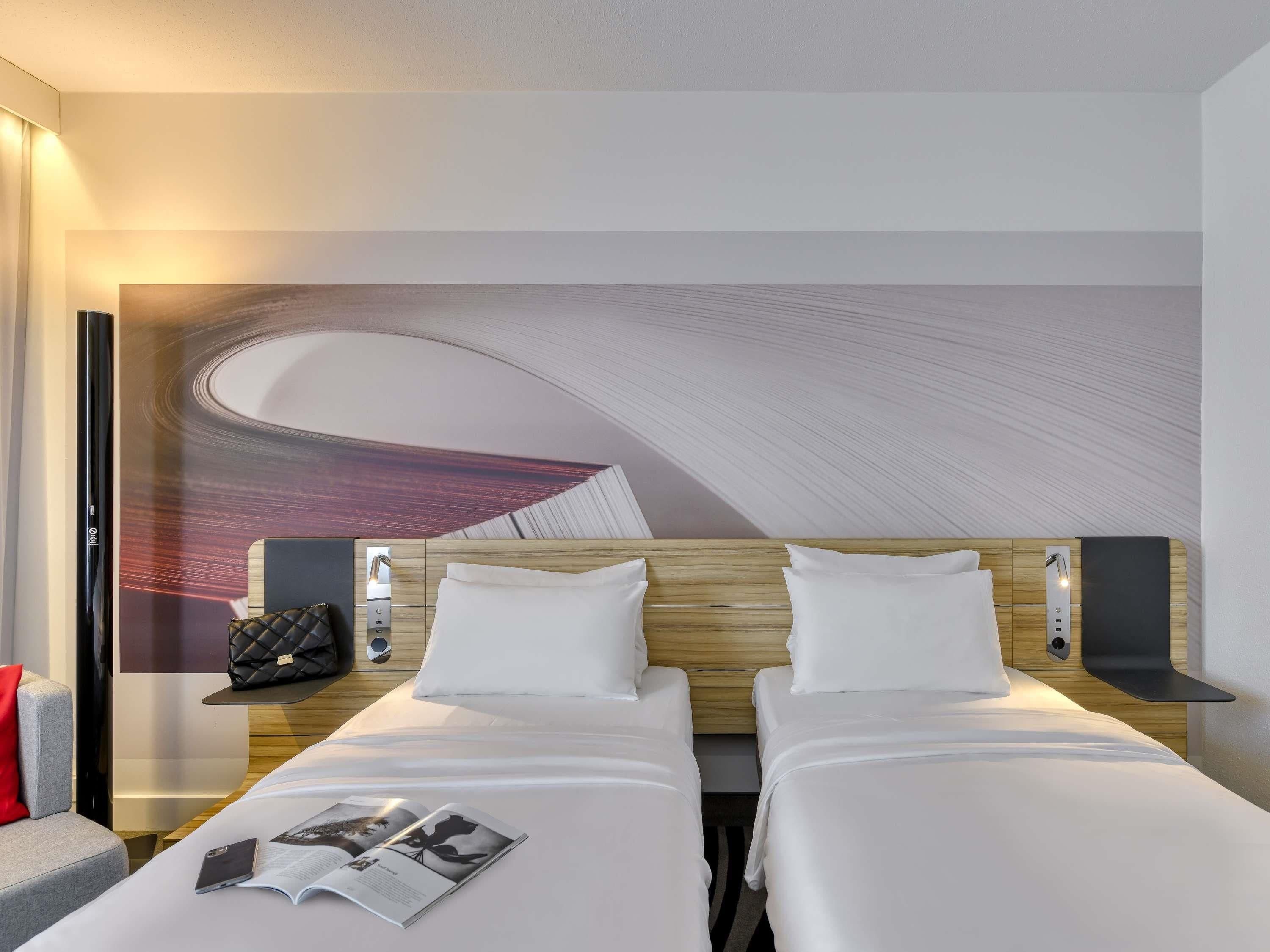 A hotel room with two beds, a large abstract mural on the wall, a small table with a handbag, and a magazine on the bed.