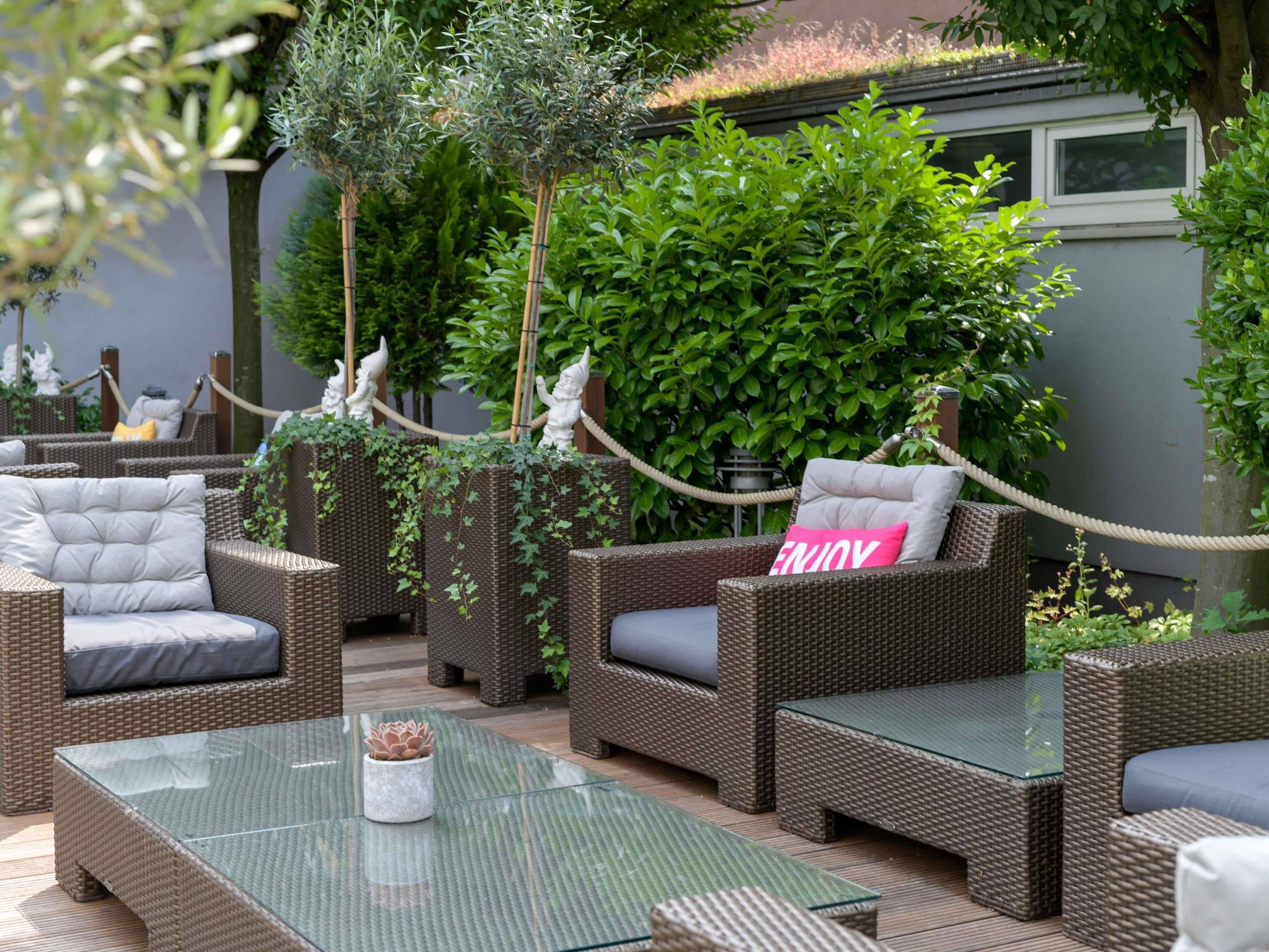 Een terras met rieten meubels, een tafel met glazen blad en groen op de achtergrond.