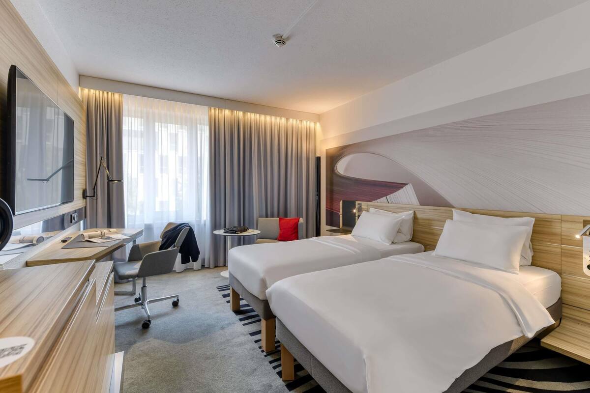 Hotellihuone, jossa on sänky, työpöytä tuolilla, televisio ja suuri ikkuna, jossa on verhot.