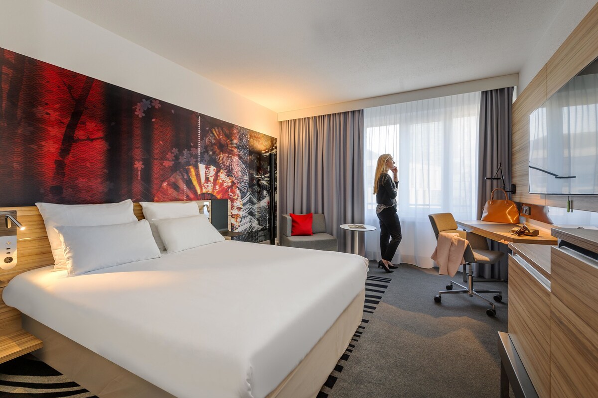 Ein Hotelzimmer mit einem großen Bett, einem Schreibtisch, einem Stuhl, einem kleinen Tisch und einem großen Fenster mit Vorhängen.