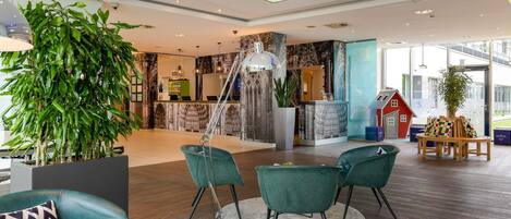 A modern lobby with a reception desk, green chairs, a large potted plant, and a view of the outside.