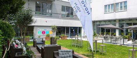 Un área de estar al aire libre con mobiliario de mimbre, una bandera y un edificio con ventanas amplias y un letrero.